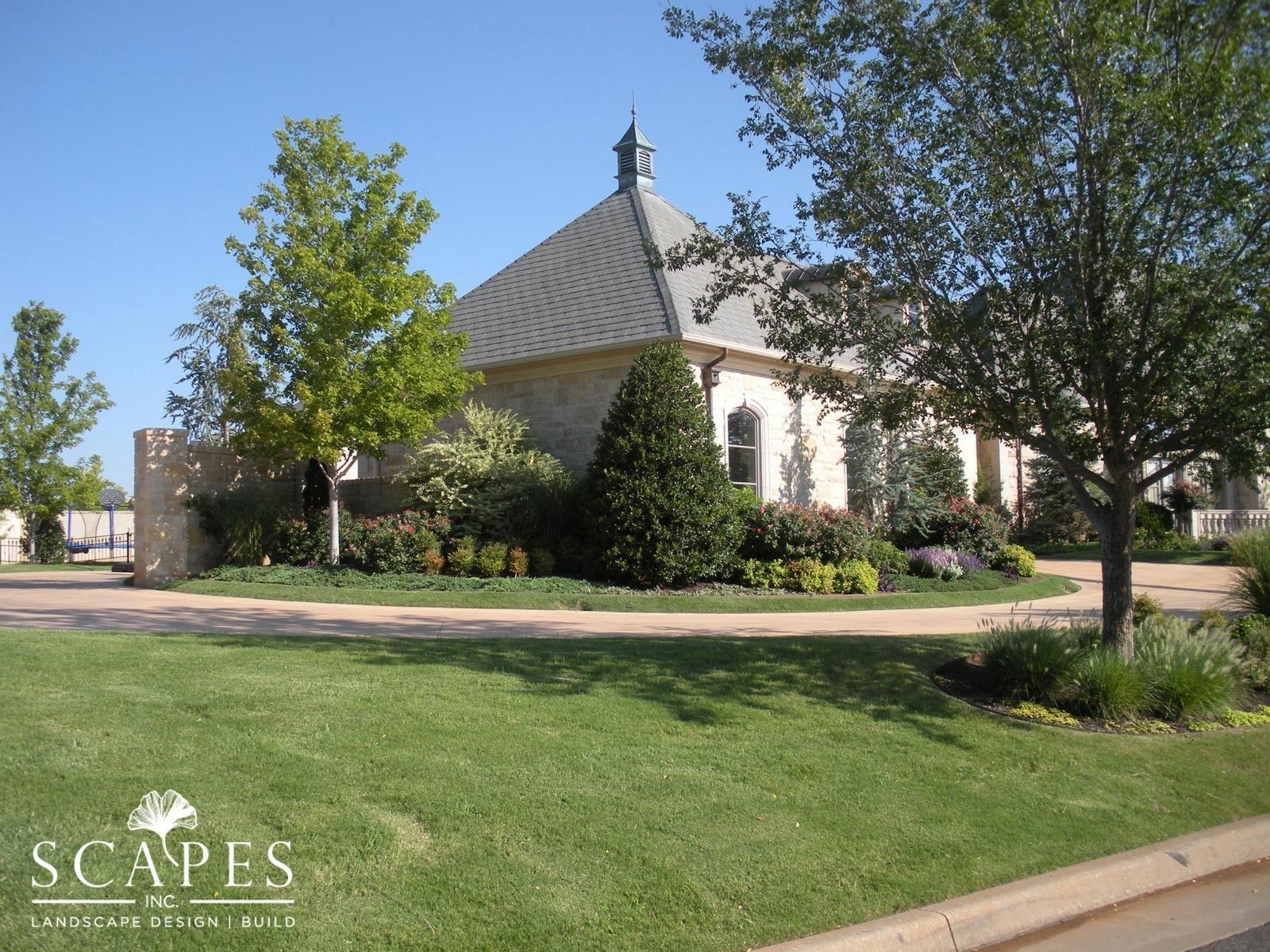 A house with a patterned roof, landscaping, and a circular driveway on a grassy lawn. A tree shades the right side.