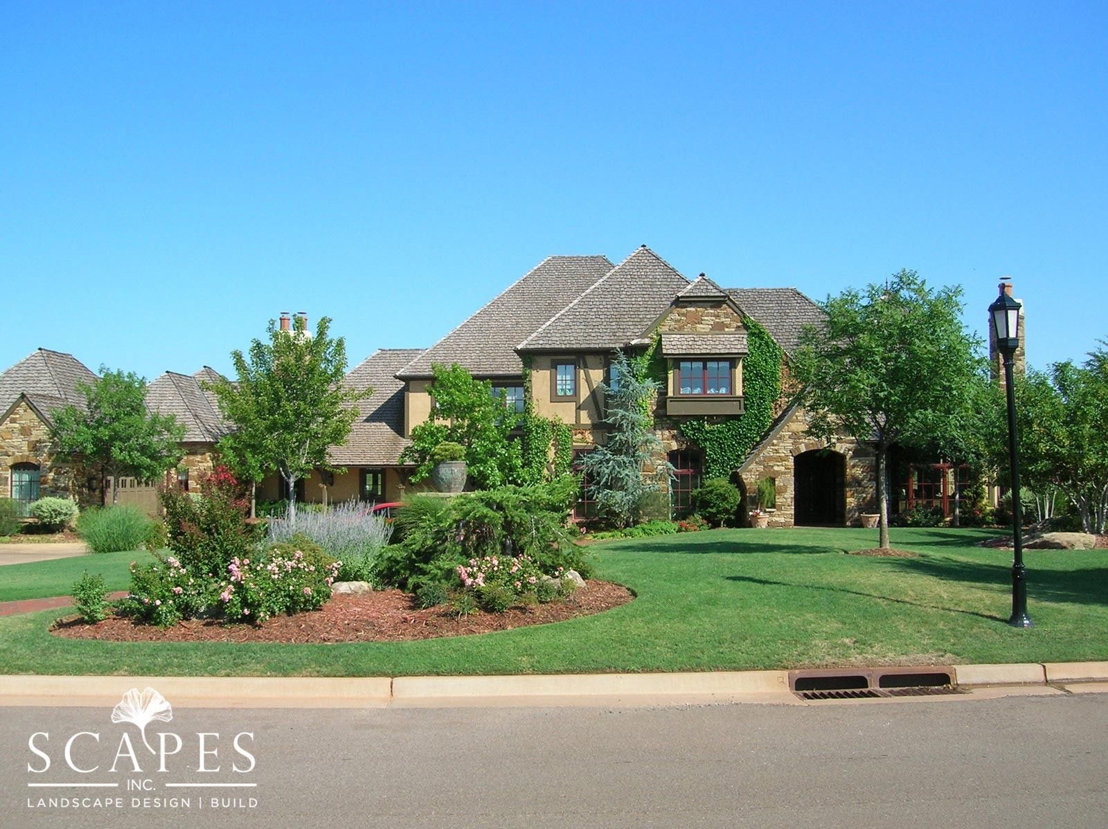 Large stone house with manicured lawn, trees, and flower beds under a clear blue sky.