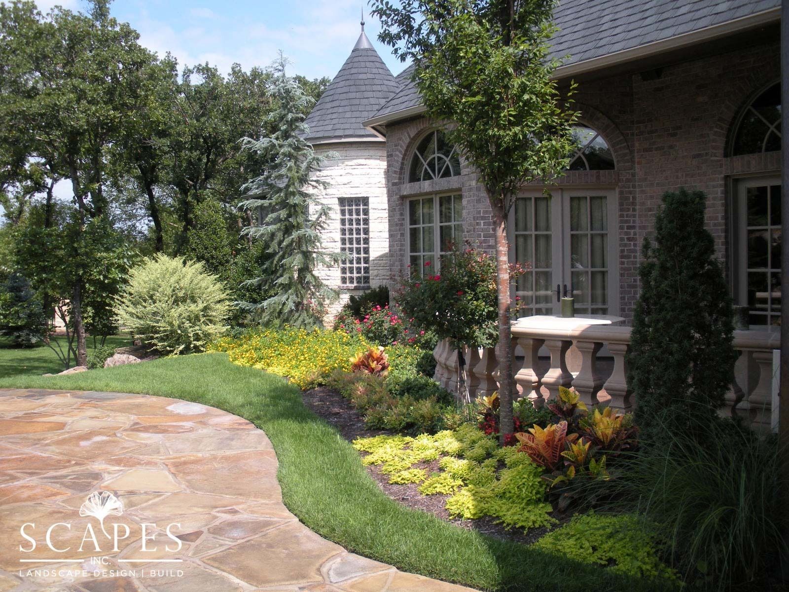 Landscaped yard with stone patio and flower beds in front of a brick house with a turret and arched windows.