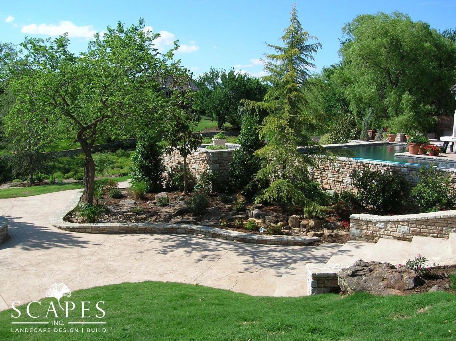 Landscaped garden with gravel paths, stone walls, various trees, and a pool area. Green grass in foreground, blue sky overhead.