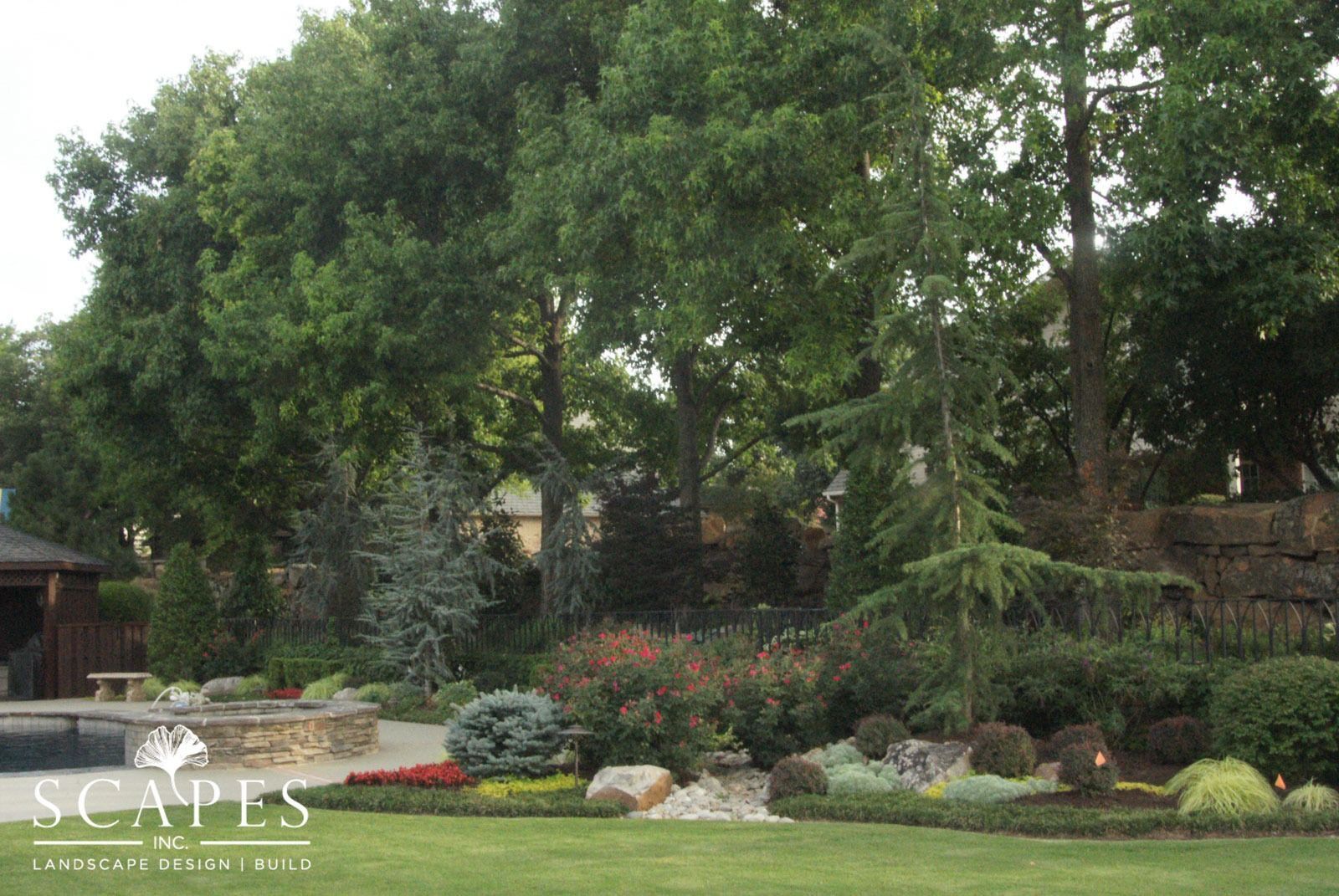Landscaped backyard with lush greenery, shrubs, and trees bordering a lawn, near a pool and stone wall.