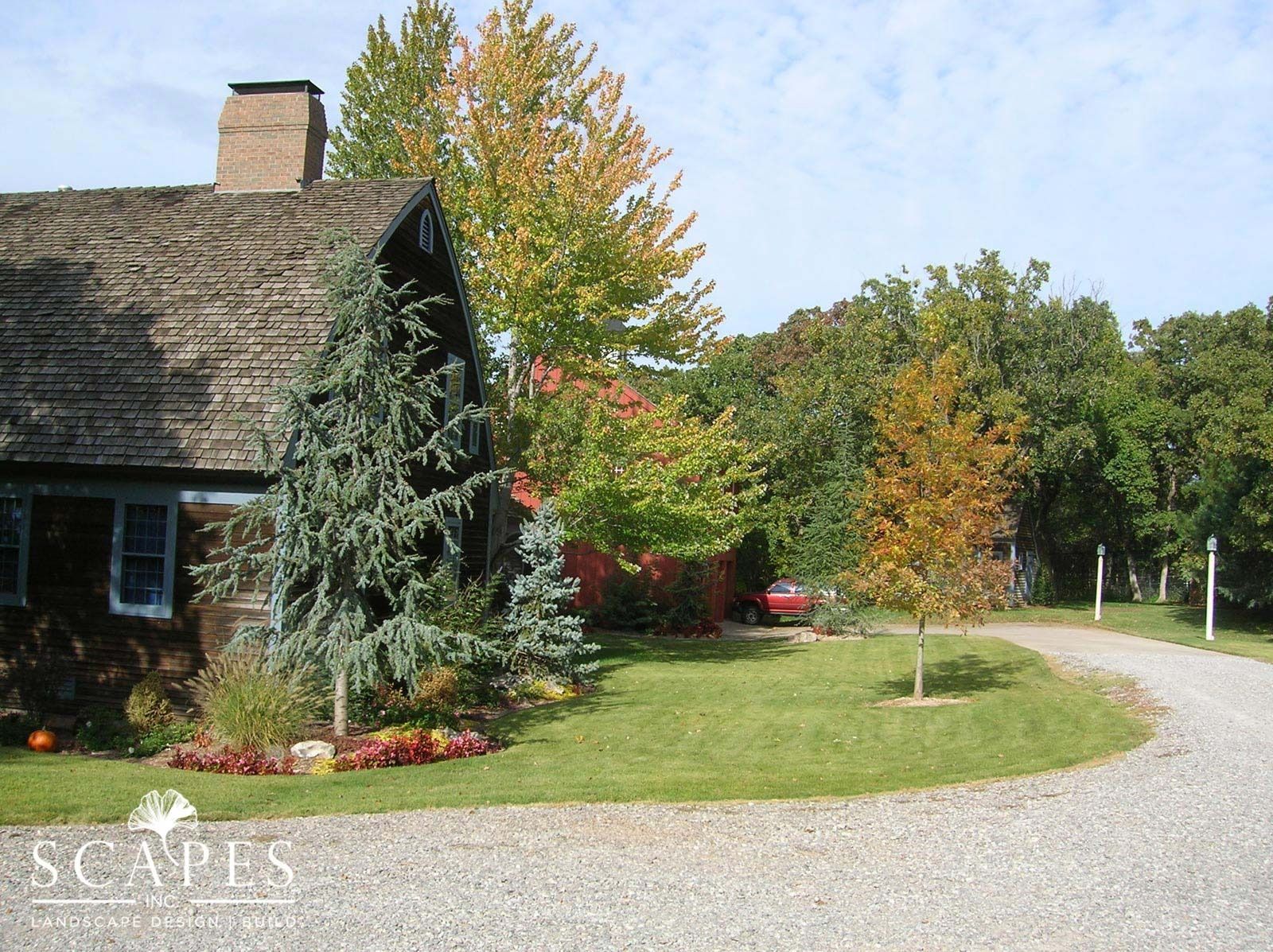 Stone house with a textured roof, a curved gravel driveway, and surrounding autumn trees. A blue spruce is in front of the house.