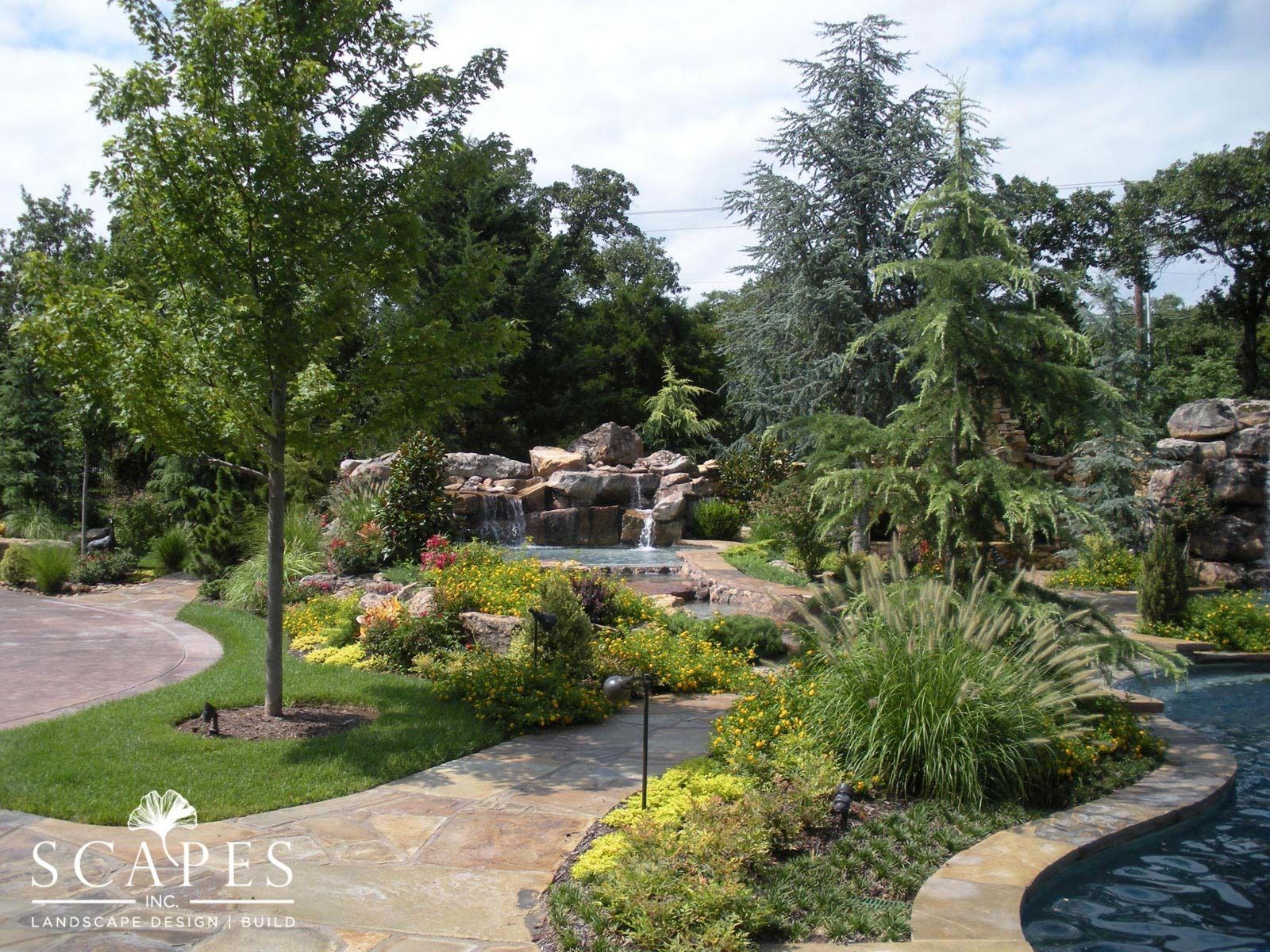 Landscaped backyard with a pool, waterfall, and various plants and trees under a partly cloudy sky.
