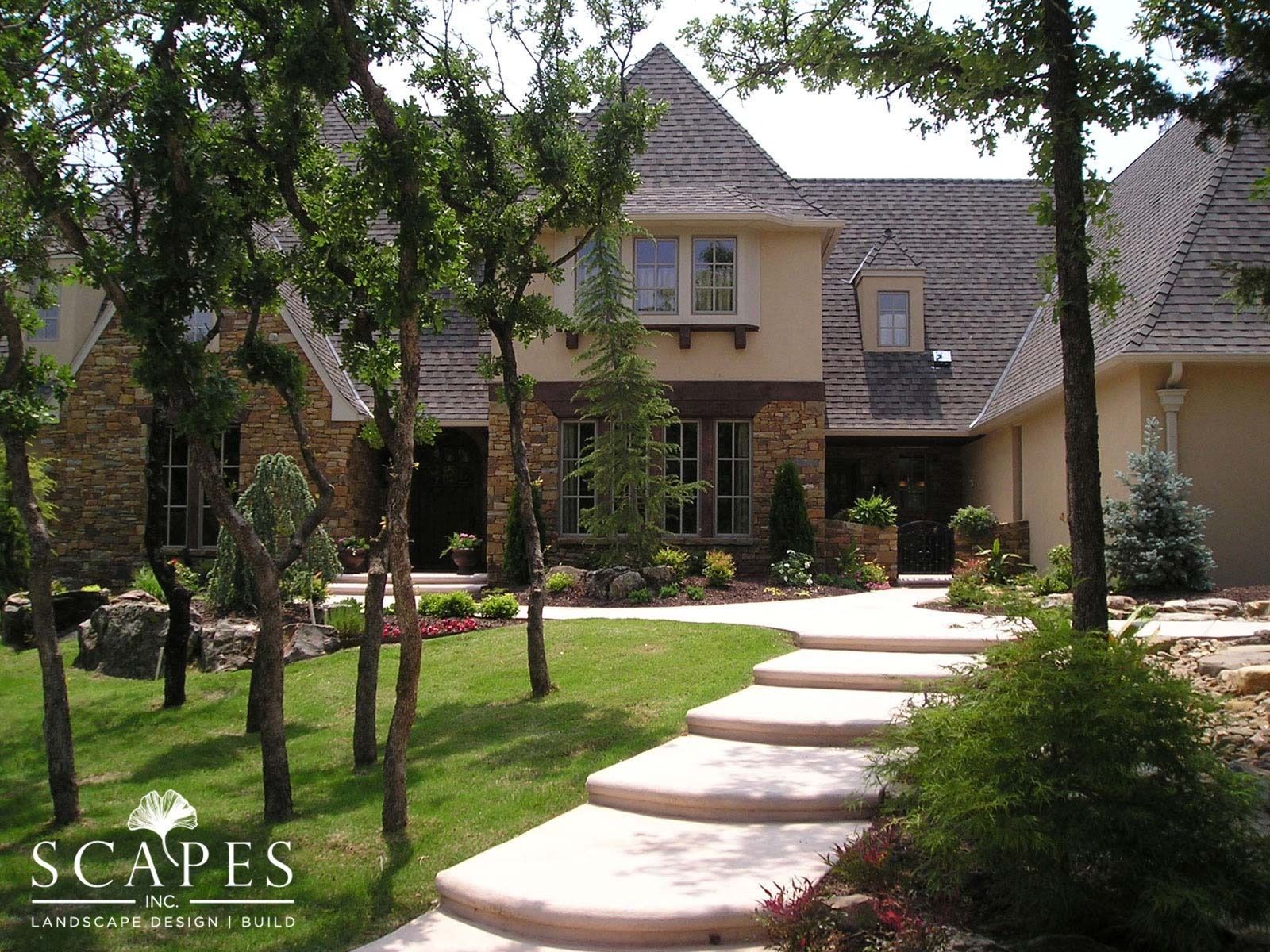 Stone and stucco house with a winding stone pathway leading to the front door; trees line the walkway, and manicured landscaping surrounds the home.