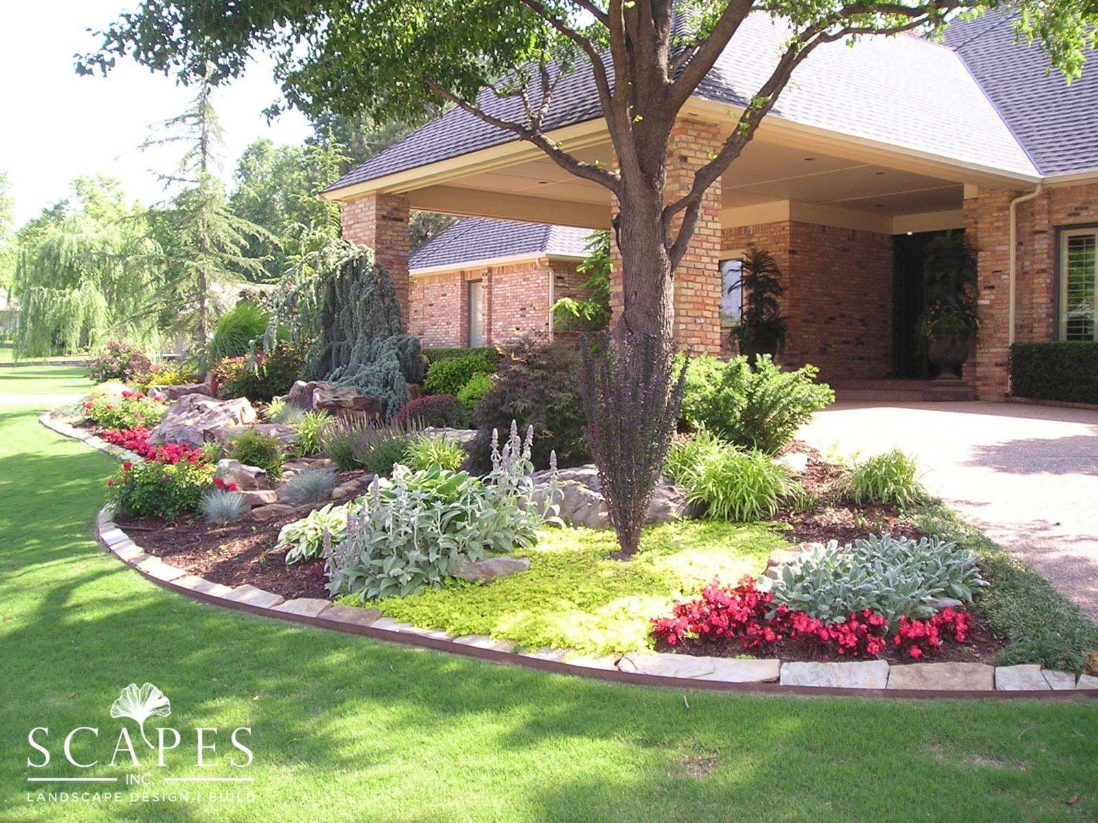 Landscaped front yard with colorful flower beds, a tree, and a brick home with a covered entryway.