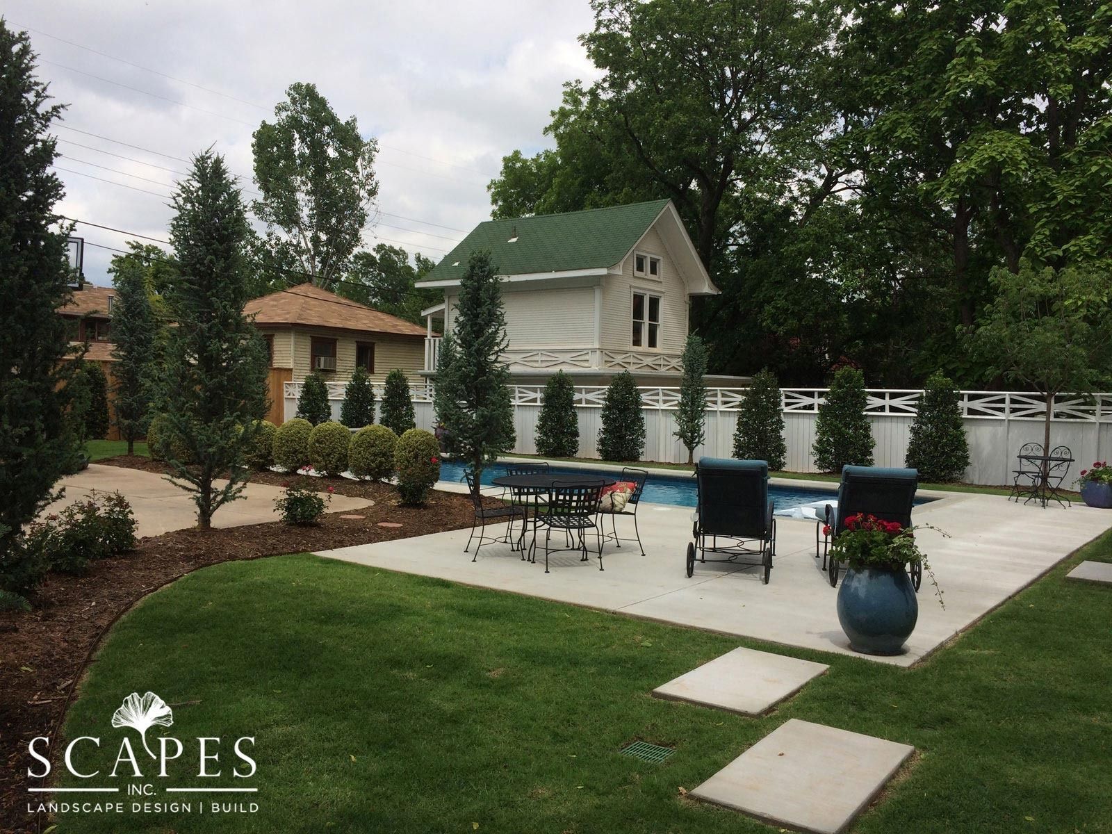 Backyard with pool and patio area, featuring a house with a green roof in the background. Landscaped with trees, bushes, and grass.