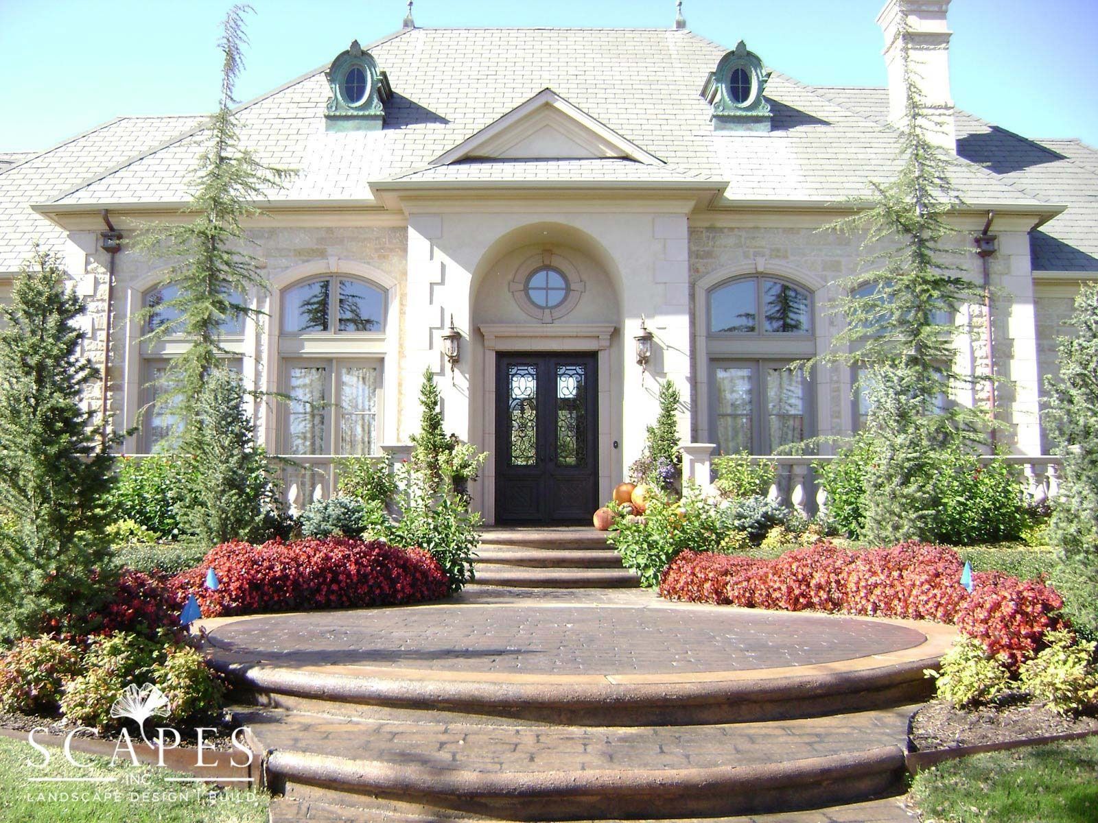 A stately beige mansion with a curved entrance and steps, featuring red and green landscaping.