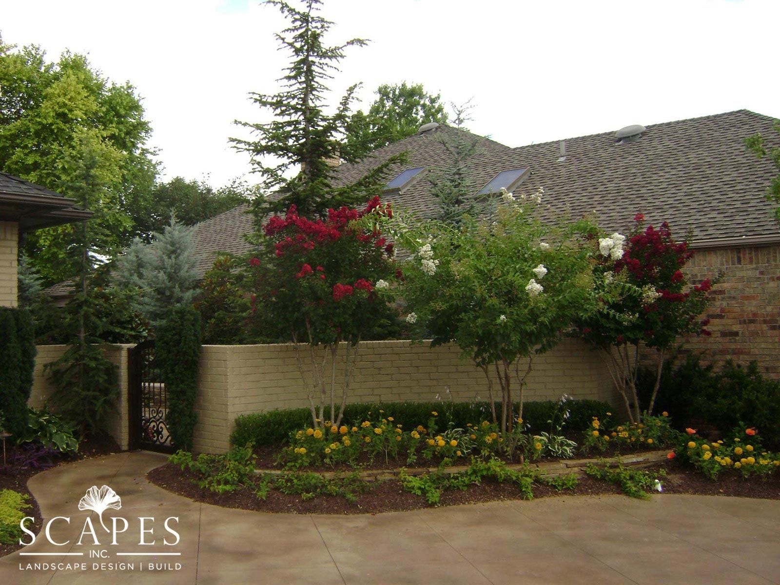 A brick home with landscaping, including trees with red and white flowers, a brick wall, and a flower bed.
