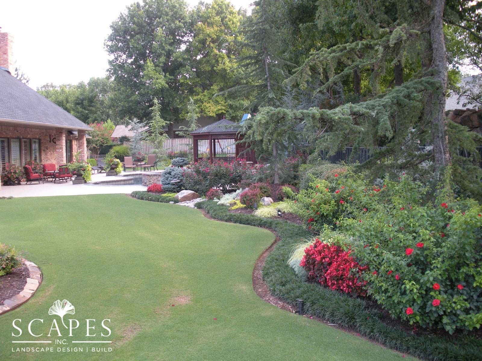 A landscaped backyard with a vibrant garden, gazebo, and brick house. Green lawn, colorful flowers, and lush trees create a serene outdoor space.