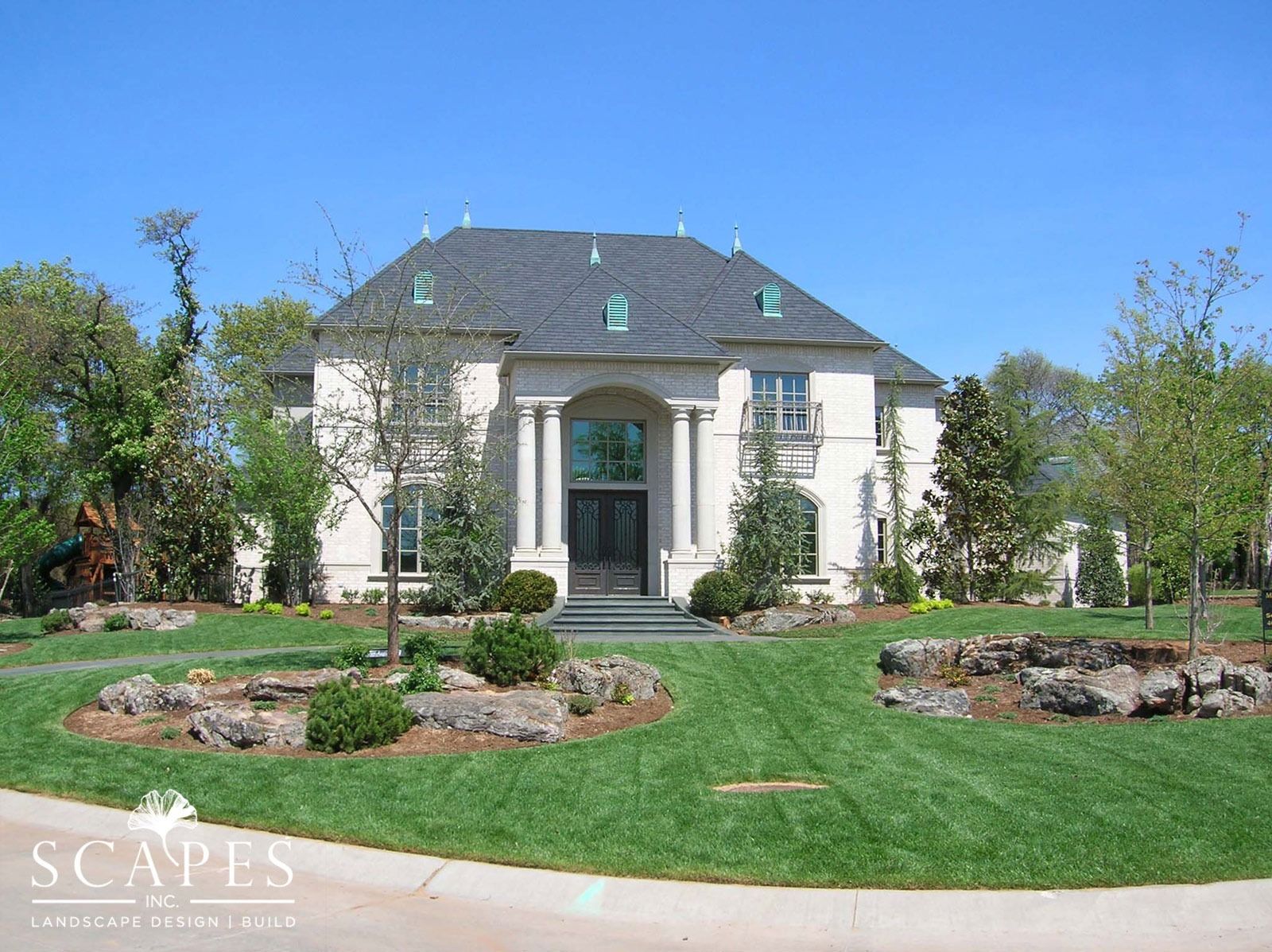 A large, white stone house with a manicured lawn and landscaping under a blue sky.