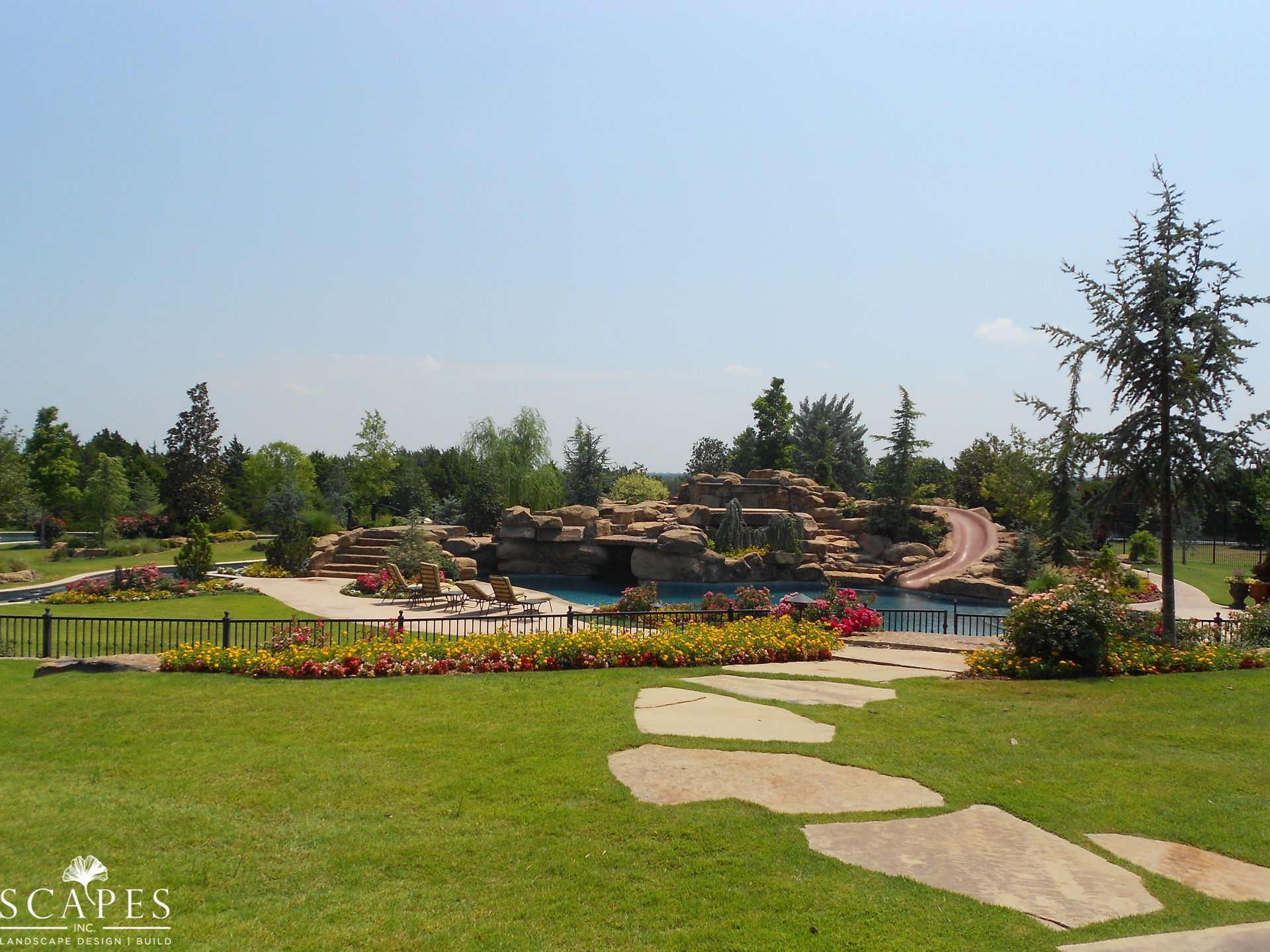 Scenic outdoor landscape with a large rock waterfall feature cascading into a pool, surrounded by greenery and a path.