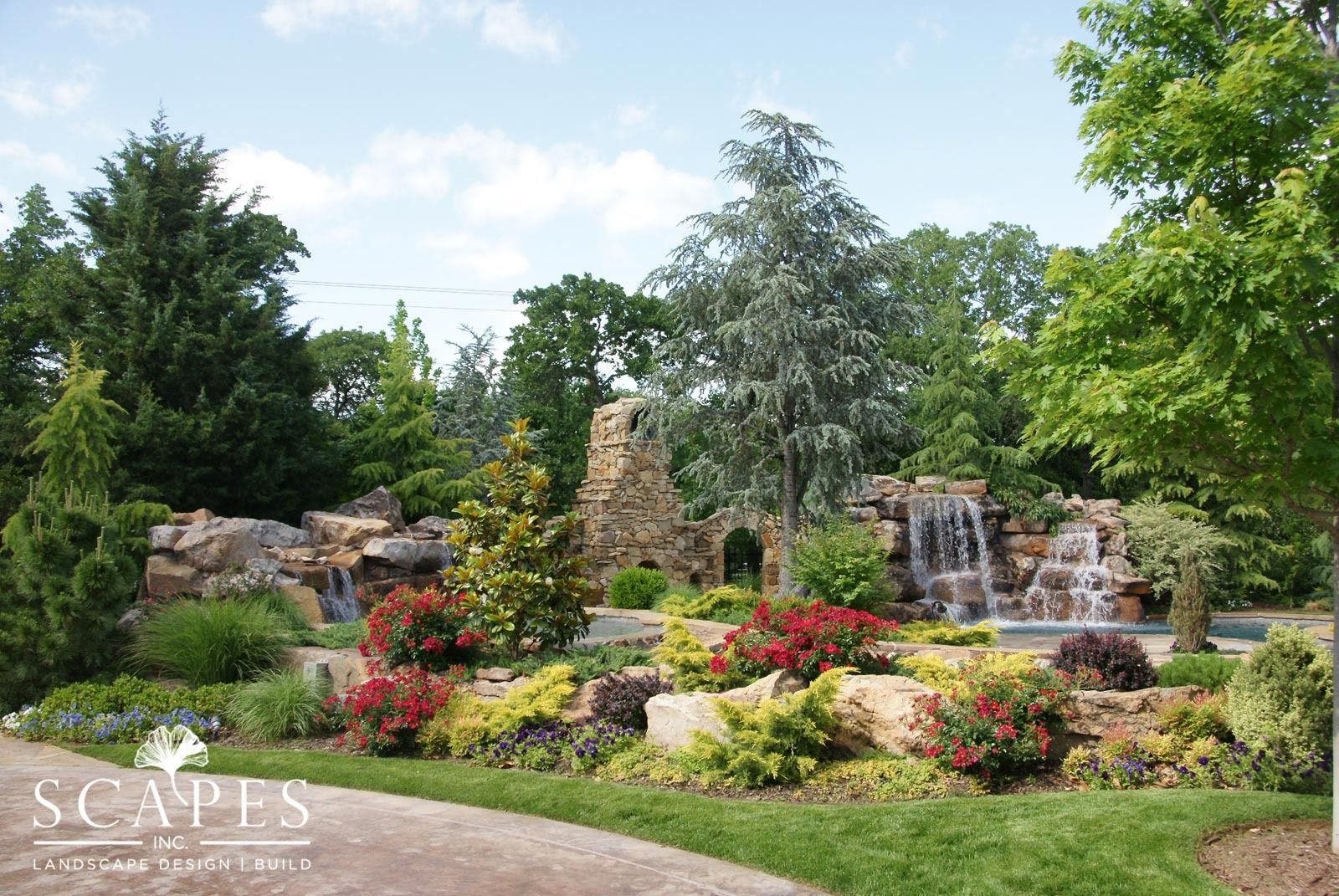 Waterfalls cascade down a rocky hillside with colorful flowers and trees in a sunny outdoor setting. A stone structure is in the center.