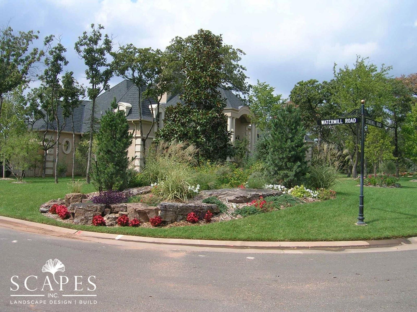 A landscaped roundabout with a house in the background. Rocks, flowers, and trees are planted within the circle of grass.