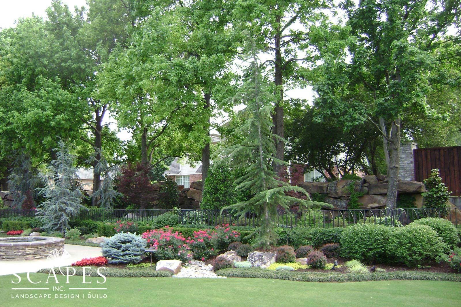 A lush landscaped garden with various trees, bushes, and colorful flowers. A stone fire pit is in the foreground, with a house visible in the distance.