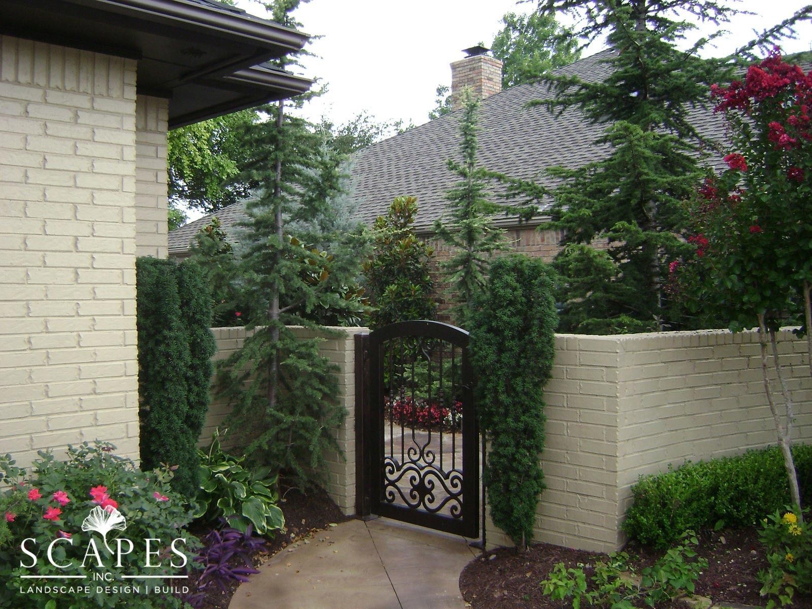 A gated entryway in a brick wall, leading to a backyard garden. Greenery and a paved path surround the decorative wrought-iron gate.