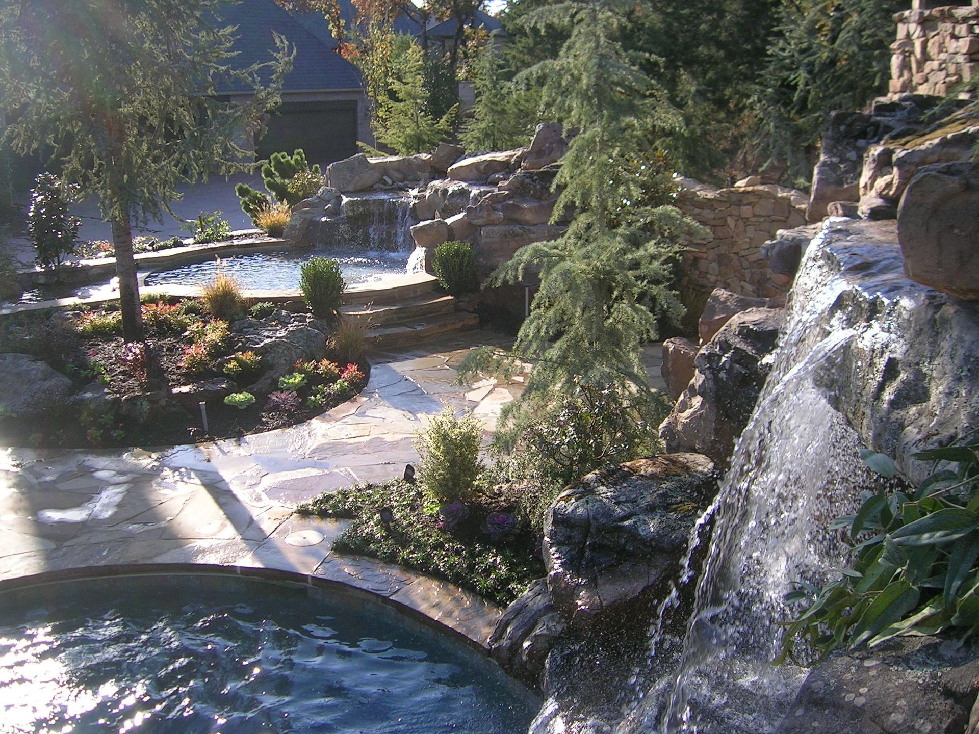 Water cascades down a rocky waterfall into a pool, with lush landscaping and another pool visible in the background.
