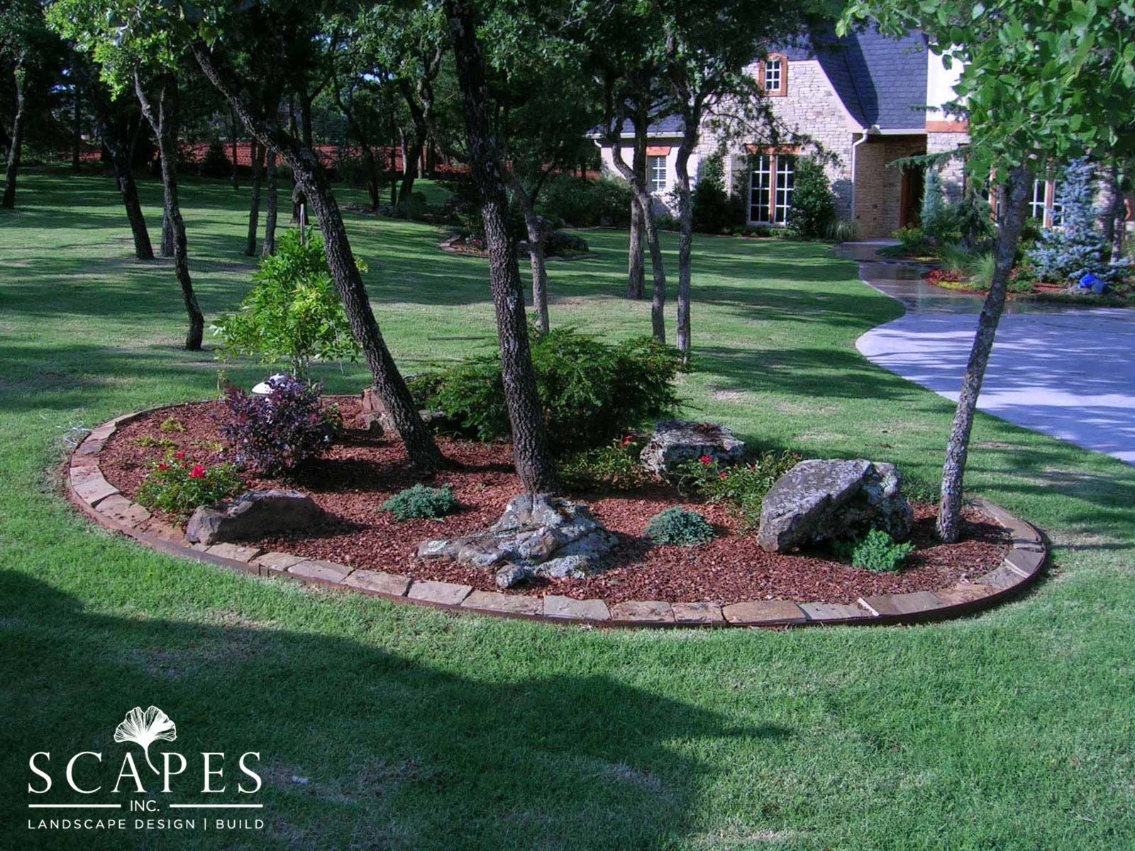 Oval-shaped garden bed in a grassy yard with trees, rocks, mulch, and colorful plants, bordered by reddish-brown pavers; a house is visible in the background.