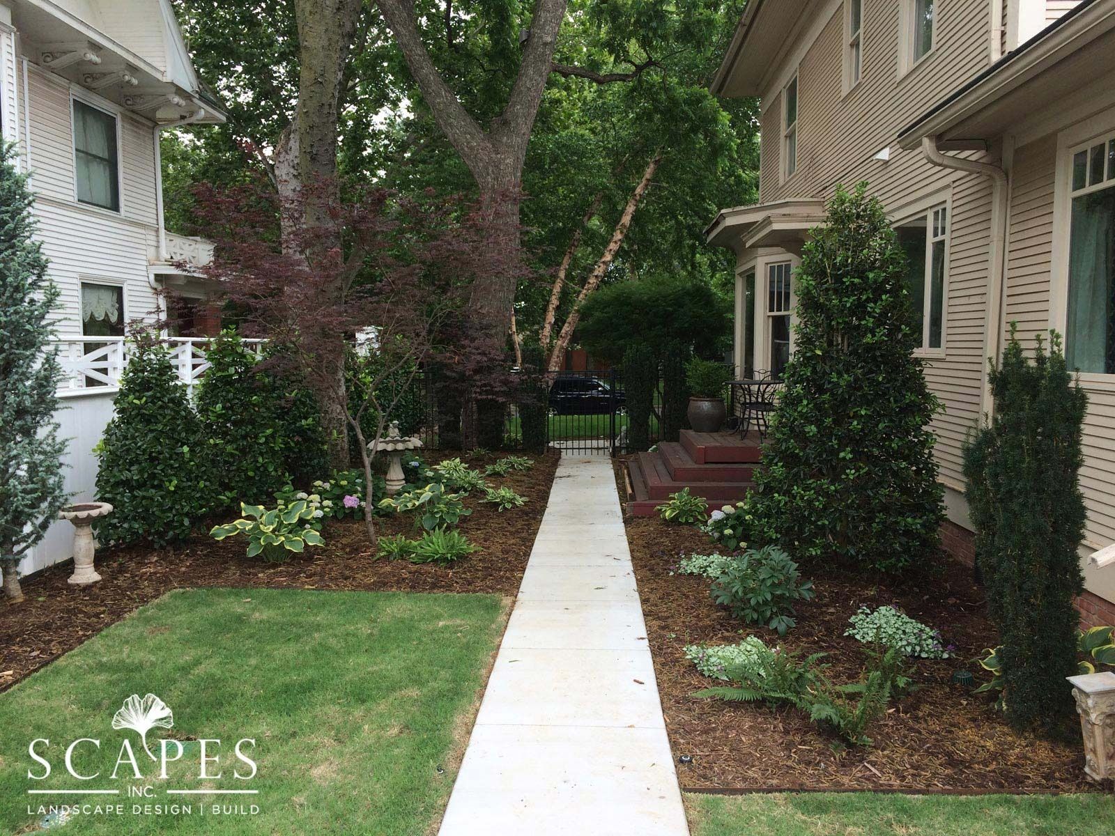 A pathway leads through a landscaped yard between two houses, with lush greenery and a small birdbath.