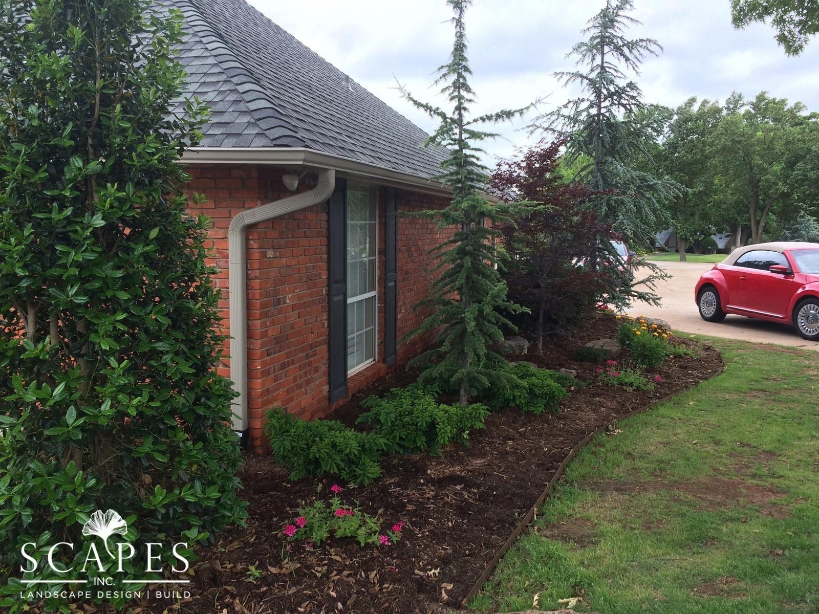 Brick house with landscaped flower bed. Green shrubs and small trees border the building and are set in dark mulch.