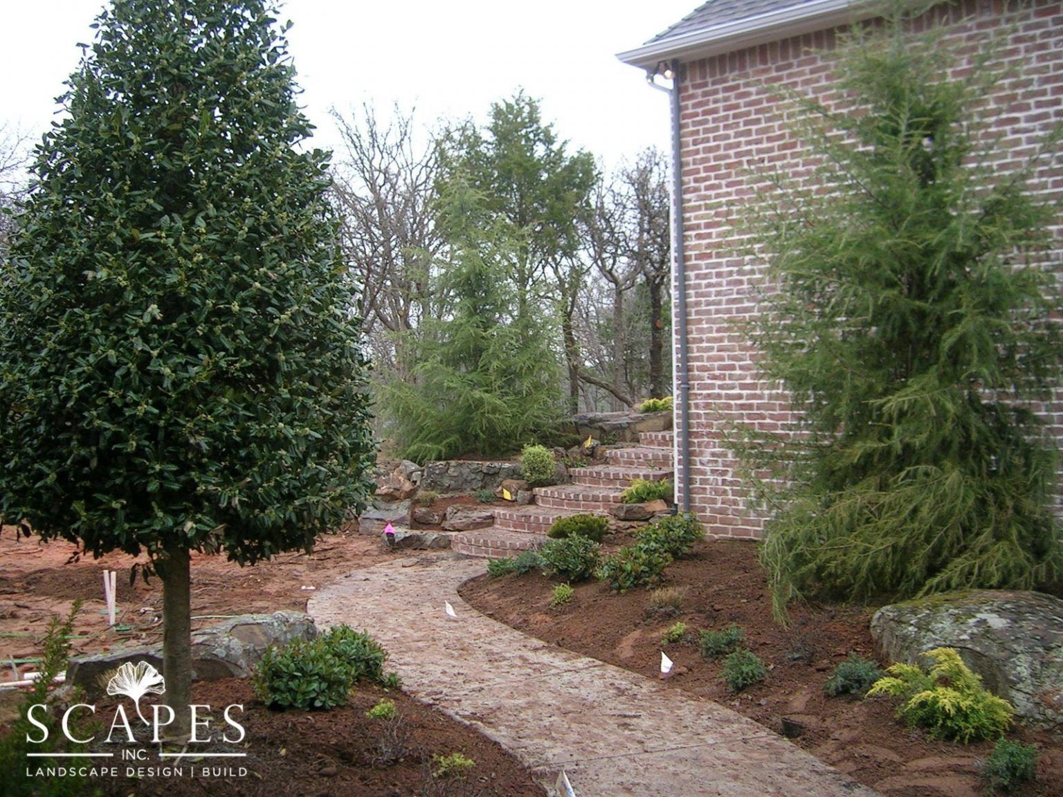 A meandering gravel path leads past various plants and a brick house with stone steps.