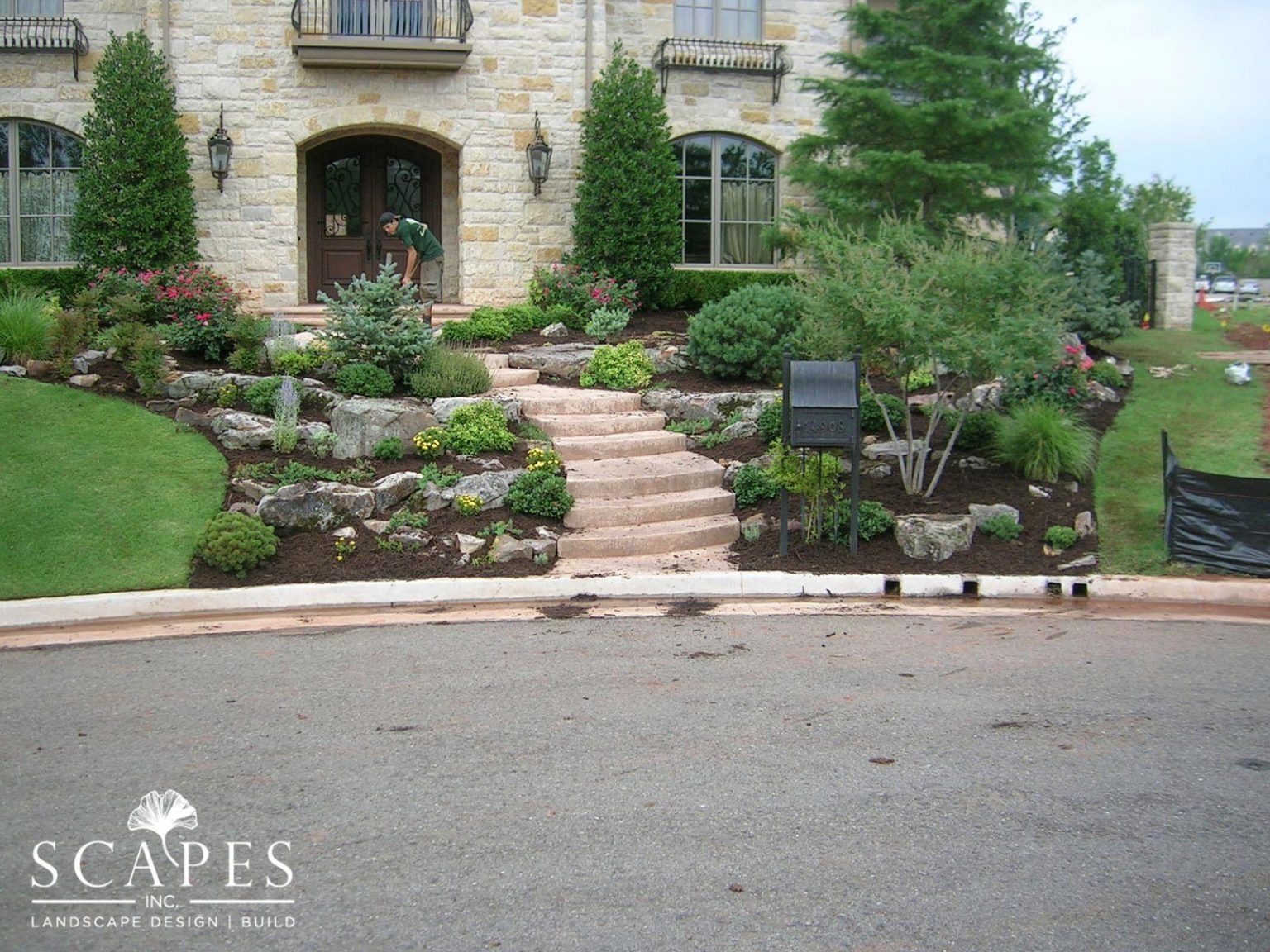 Stone steps leading up to the front door of a beige stone house with landscaped gardens, including shrubs, flowers, and trees.