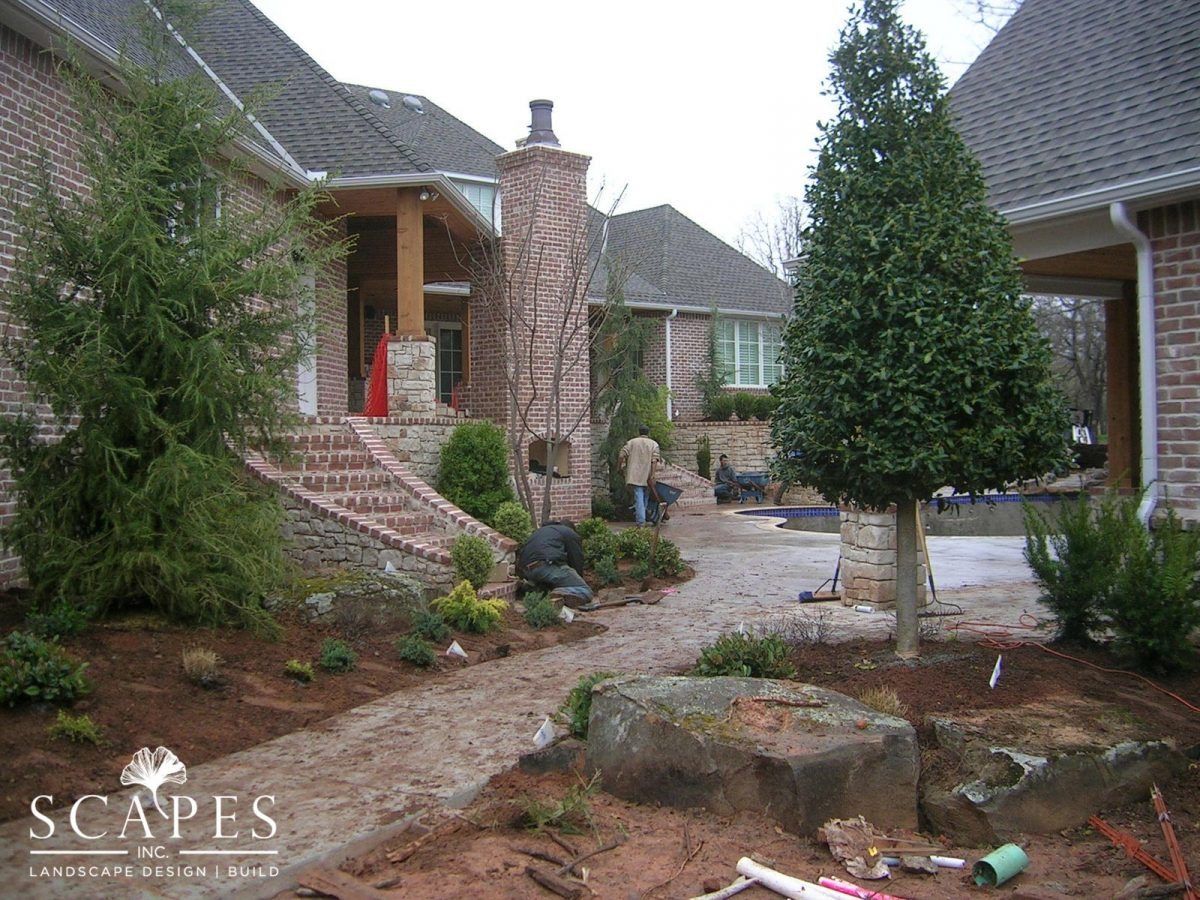 A landscaping project in progress outside a brick home. Two workers are near a gravel pathway, surrounded by newly planted trees and mulch.