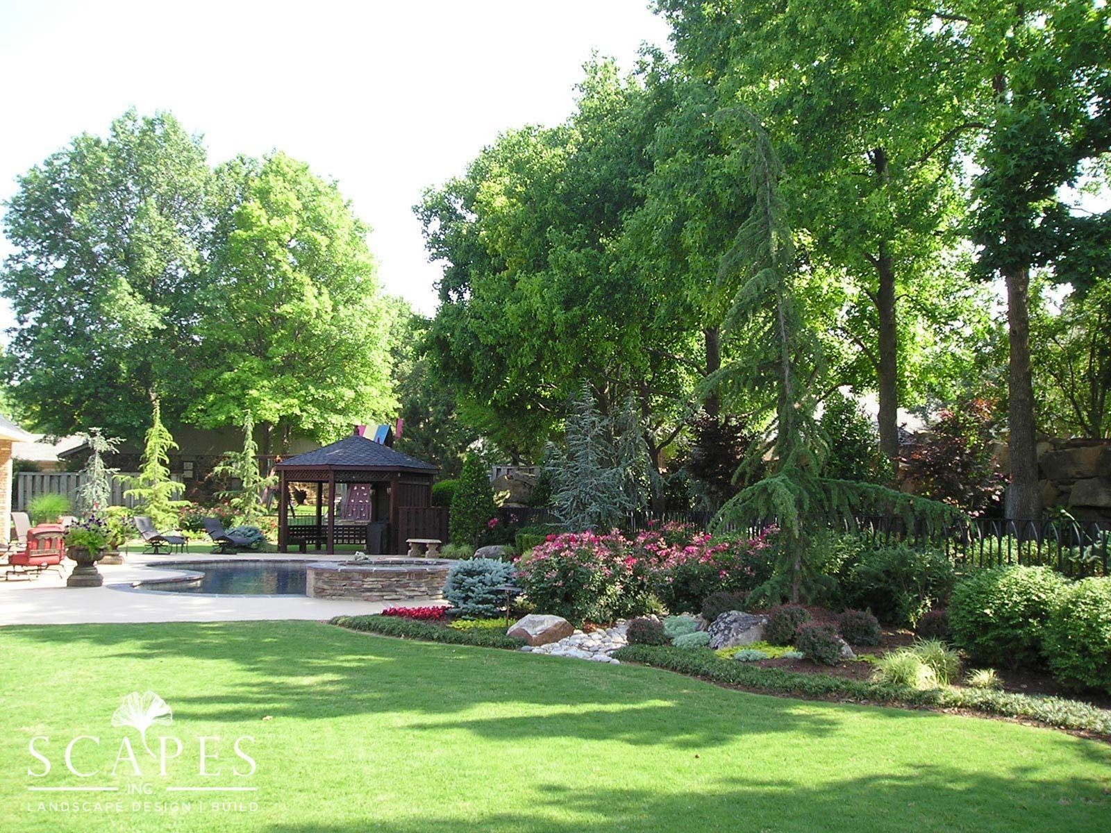 A lush, green backyard with a garden bed of colorful flowers, a gazebo, and a pool surrounded by trees under a sunny sky.