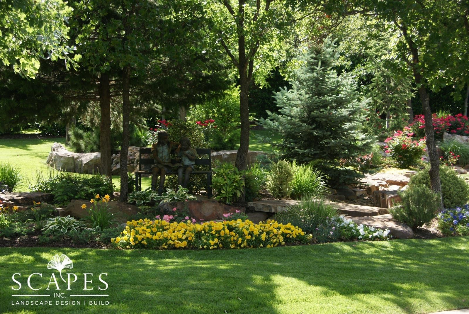 Lush garden with yellow and pink flowers, a water feature, and trees under a sunny sky.