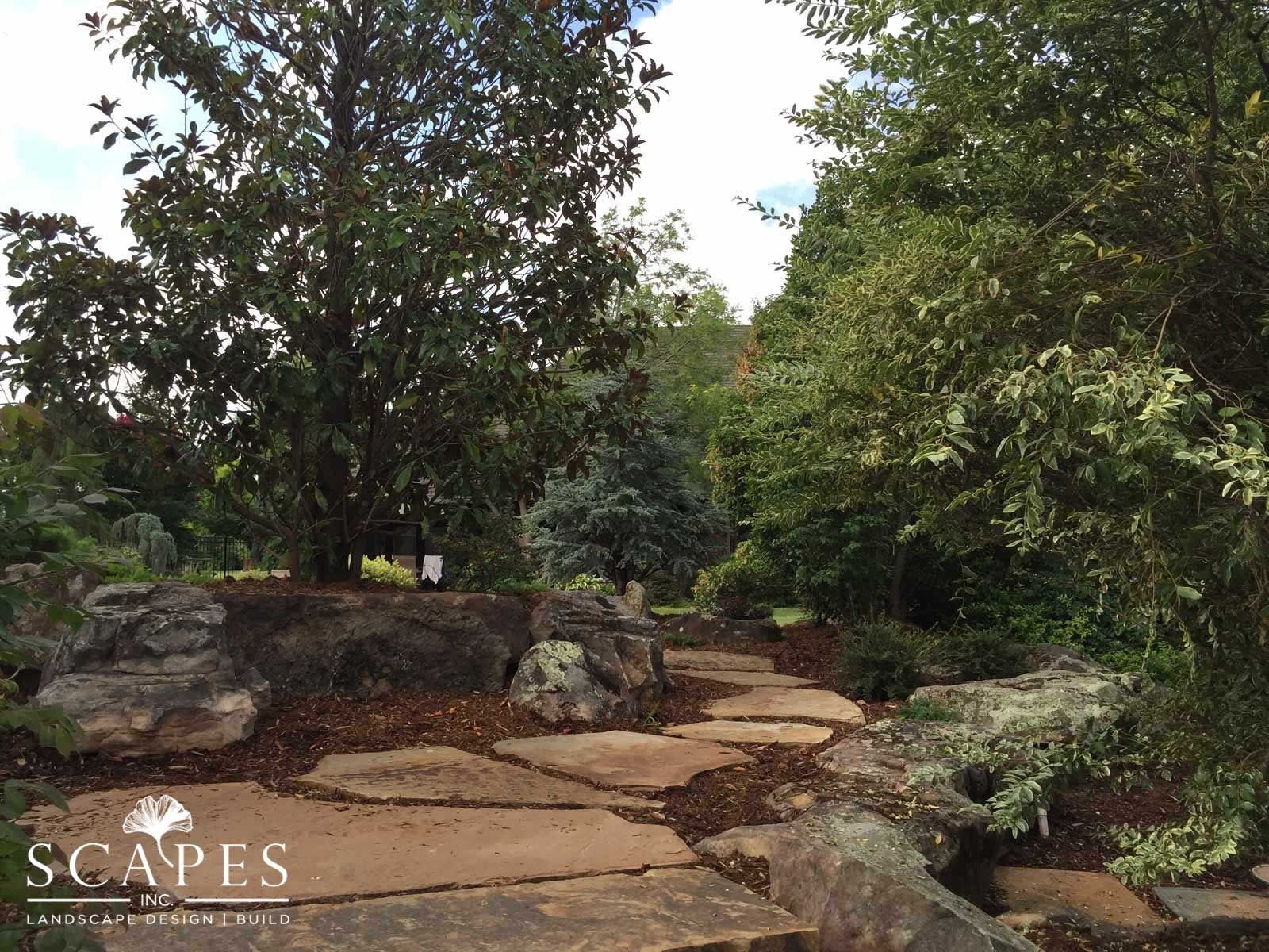 Stone path winds through a lush garden with large trees and rocks.