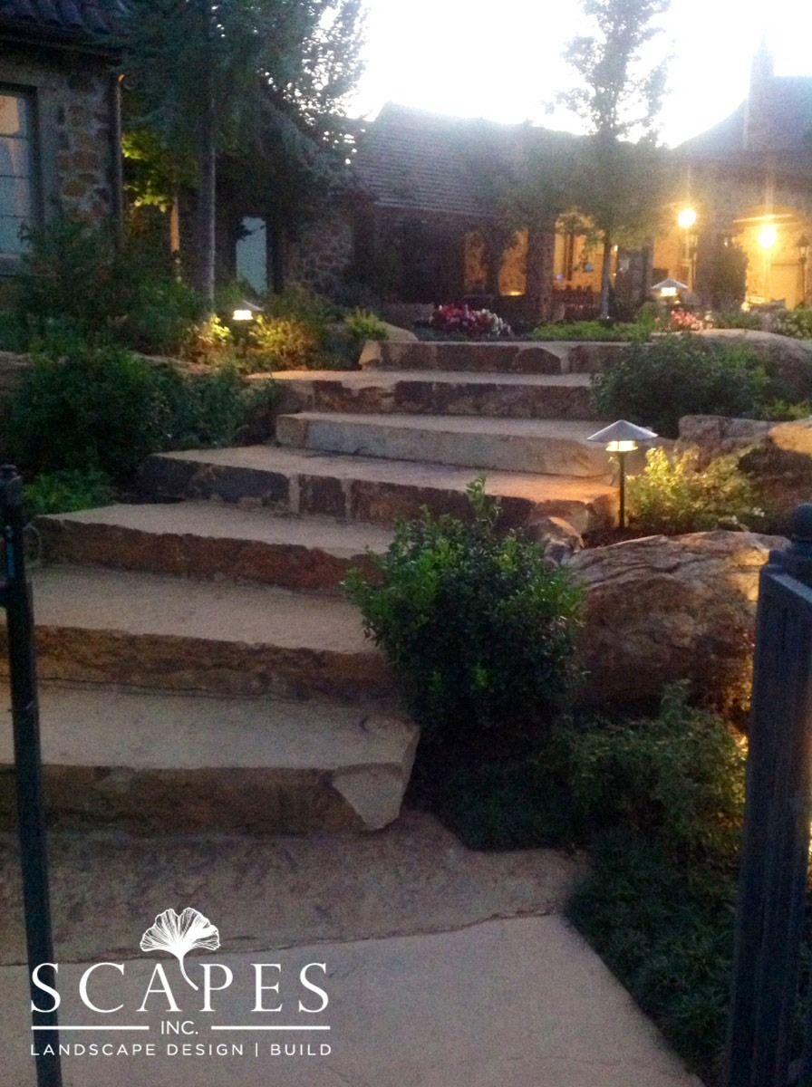 Stone steps lead up to a house, illuminated by outdoor lighting. Bushes and rocks line the path, while a paved area is in the foreground.