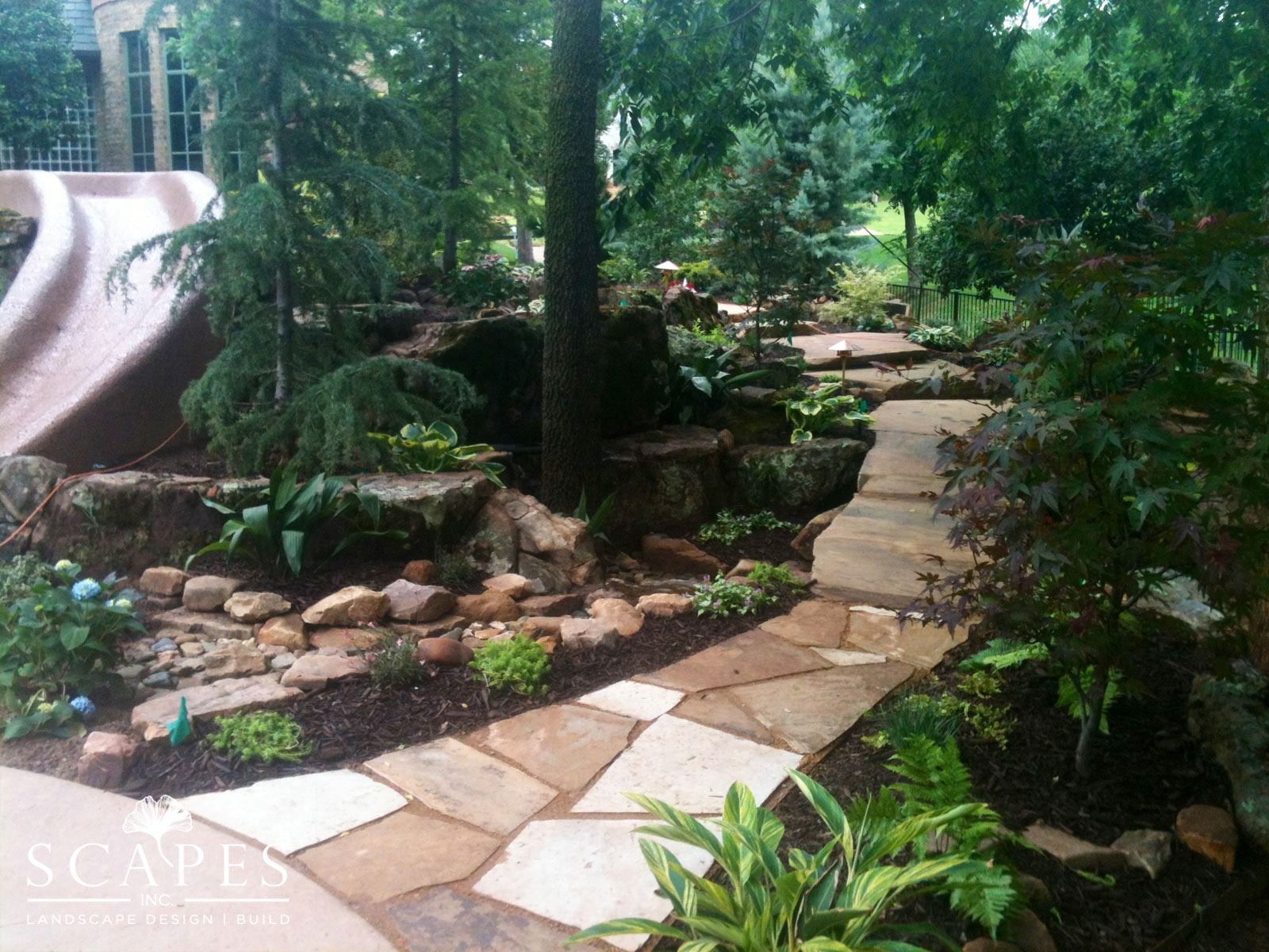 A flagstone pathway winds through a landscaped garden with rocks, trees, and lush greenery, next to a large slide.