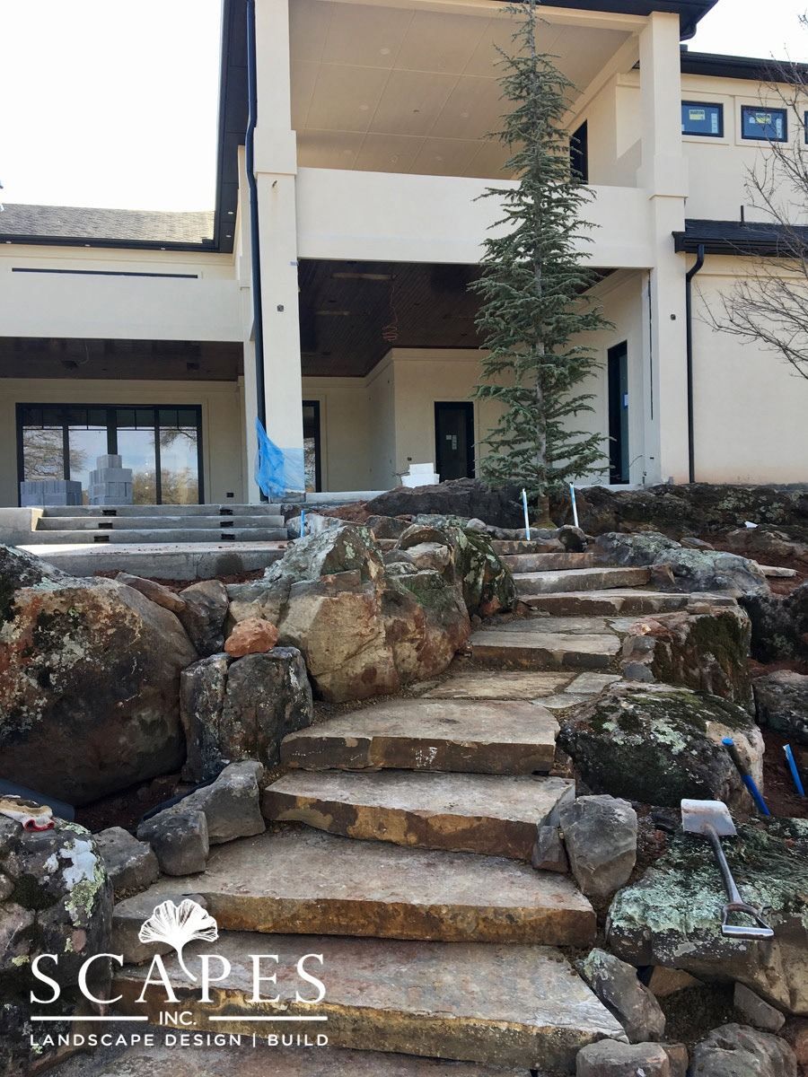 Stone steps leading up to a beige house with a balcony and black trim, surrounded by large rocks and a tree.