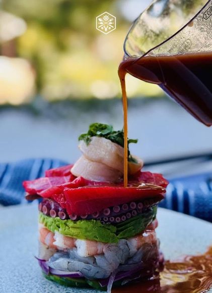 Vertical layered seafood appetizer with octopus, avocado, and salmon, topped with sauce being poured over it