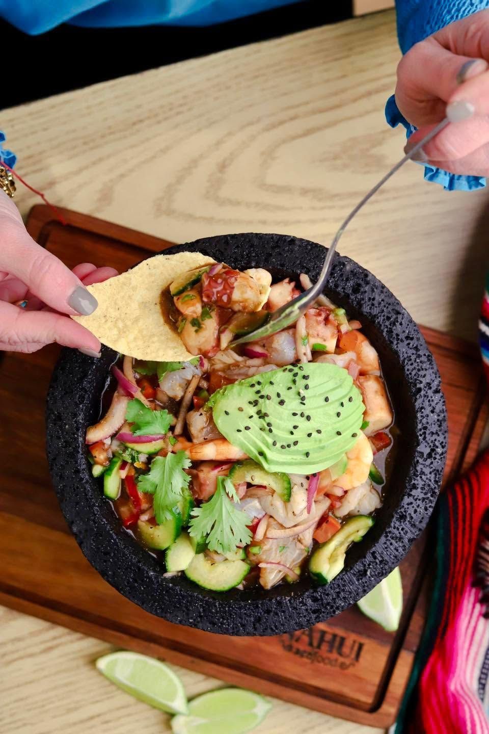 Taco bowl with avocado and salsa in a molcajete, with hands adding chips over a wooden table