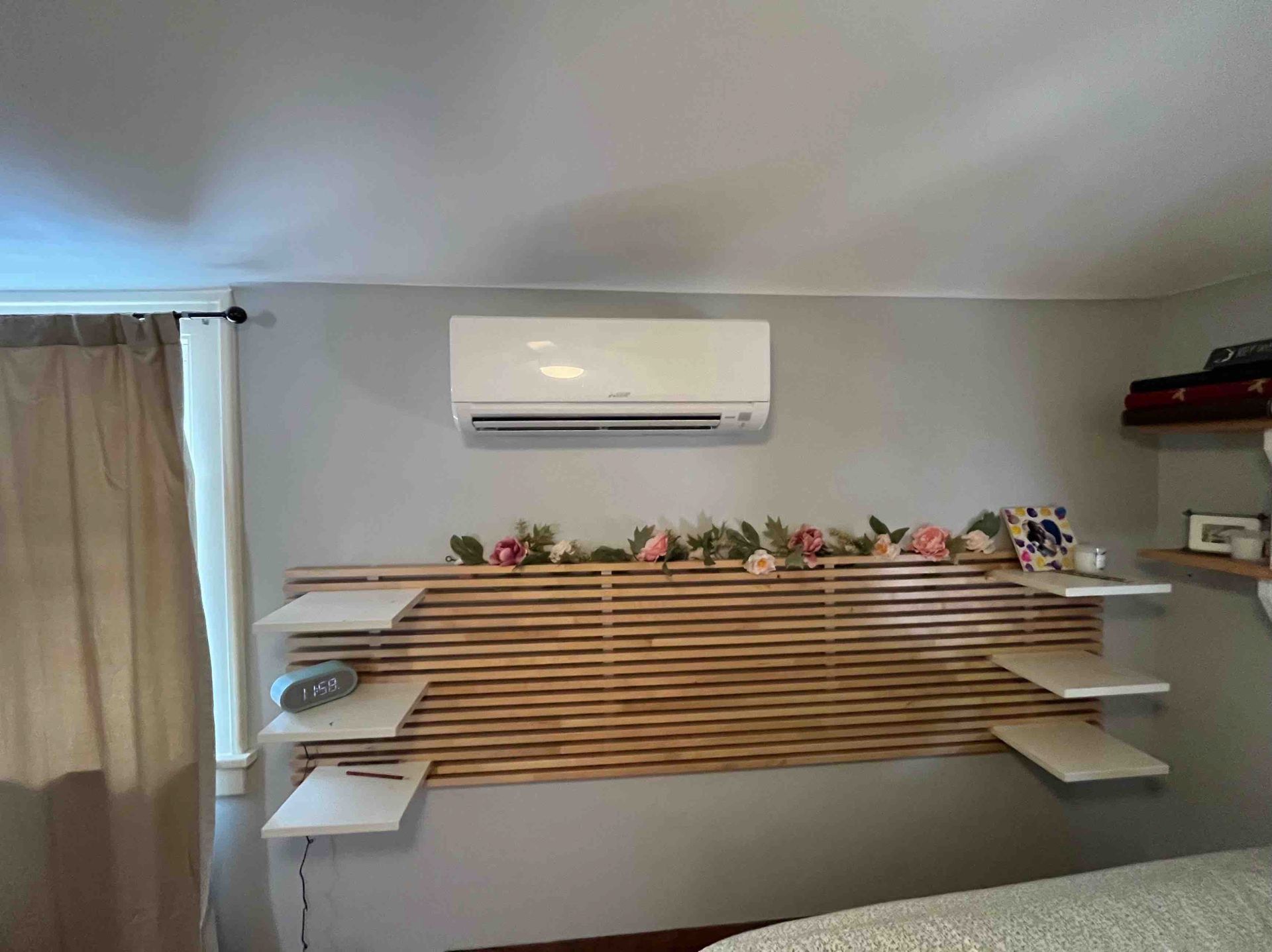 Air conditioner above a wooden headboard with floating shelves, decorated with flowers.