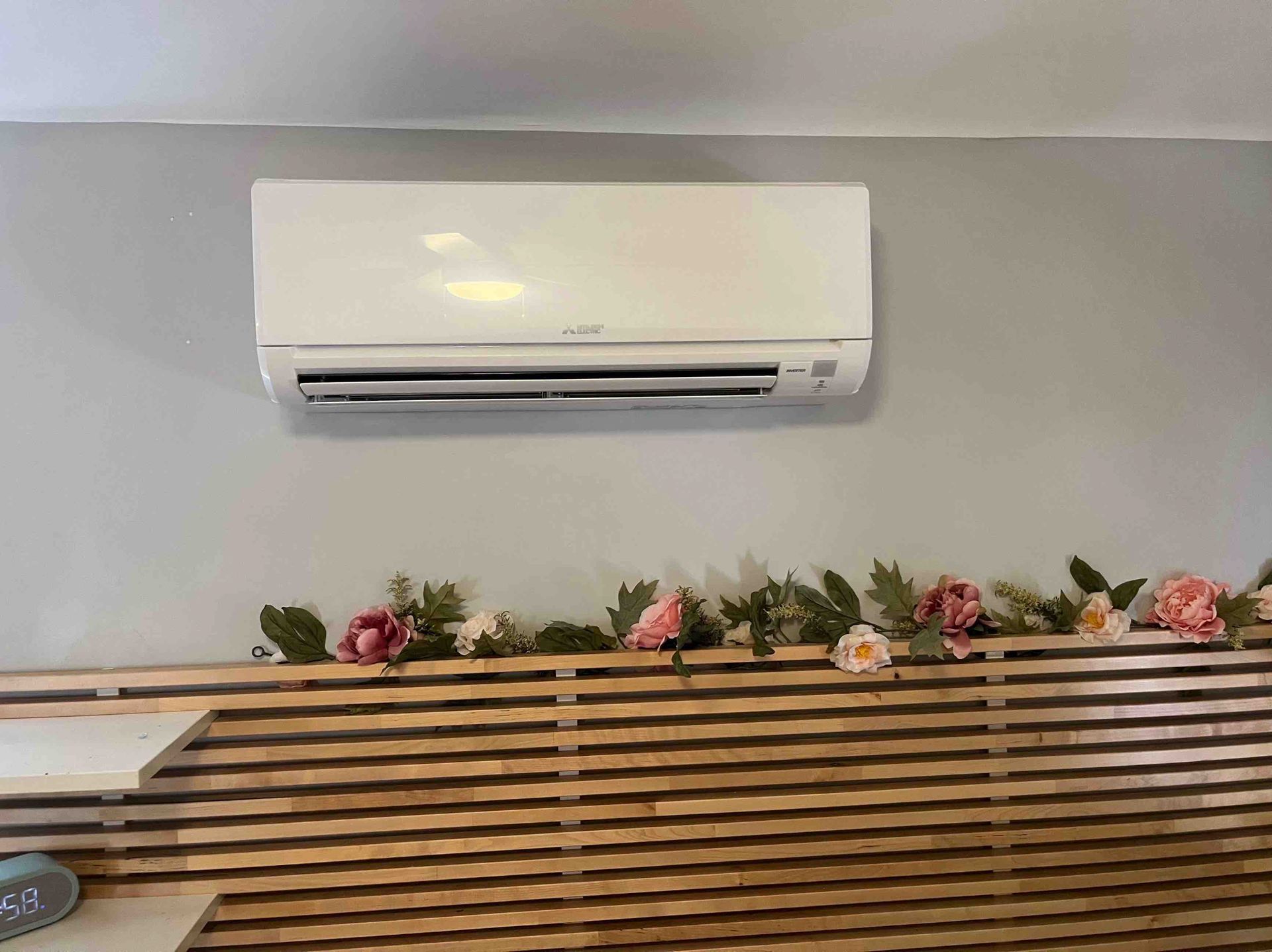 White air conditioner unit mounted on a grey wall above a wooden slat headboard decorated with flowers.
