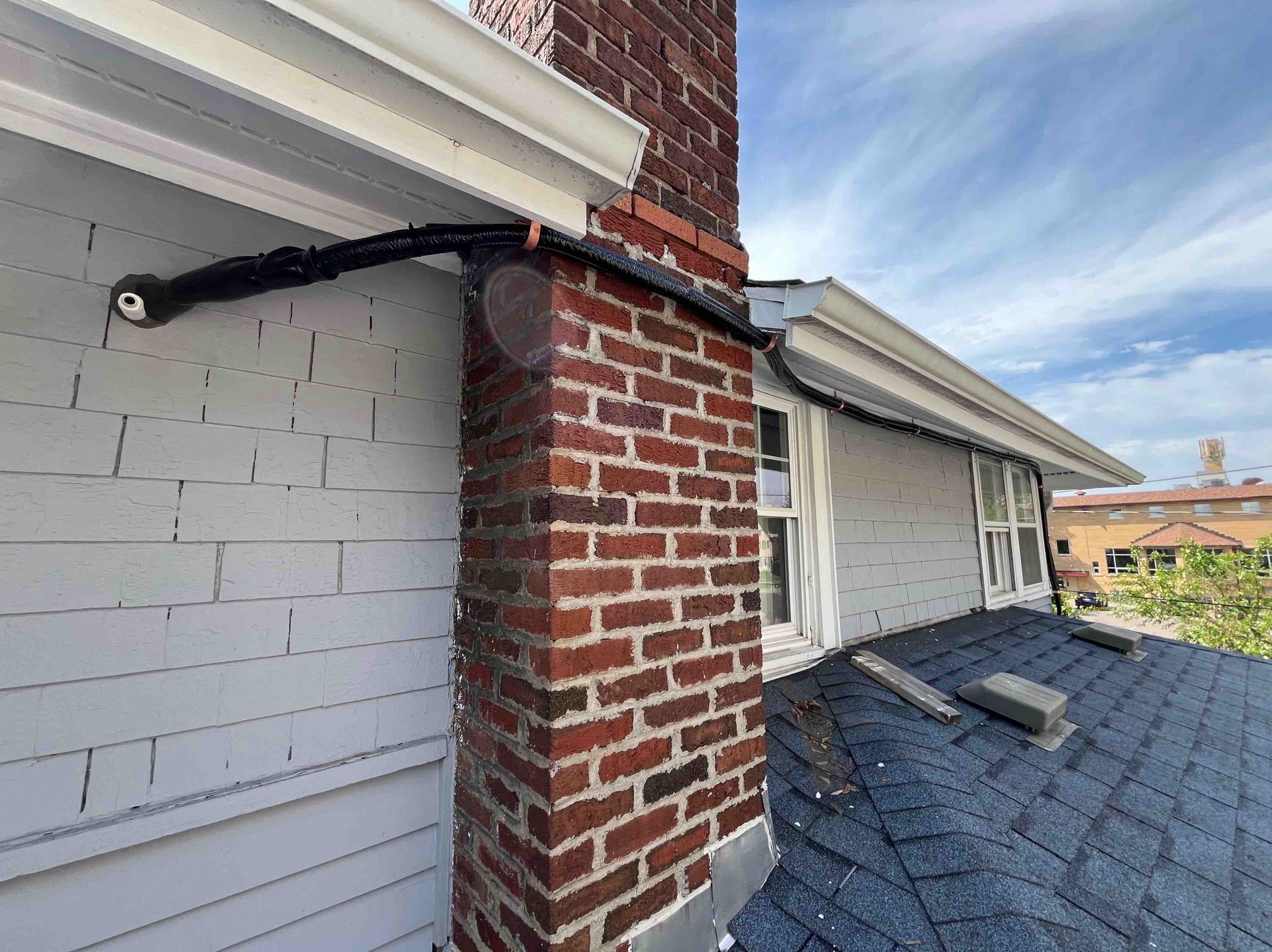 A brick chimney with black conduit, white siding, a shingled roof against a cloudy sky.