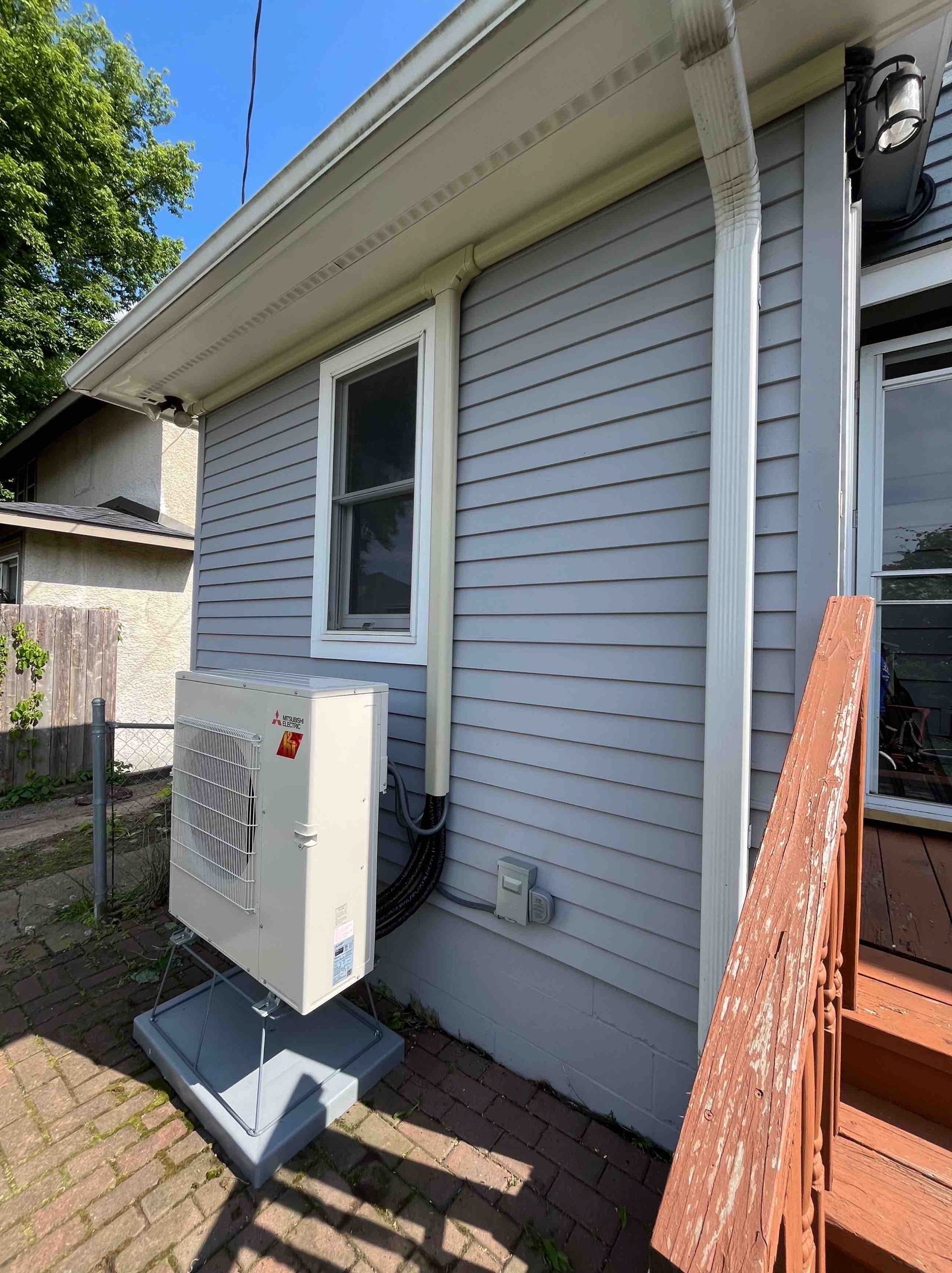 An air conditioning unit outside a gray-sided house, near a window and stairs.