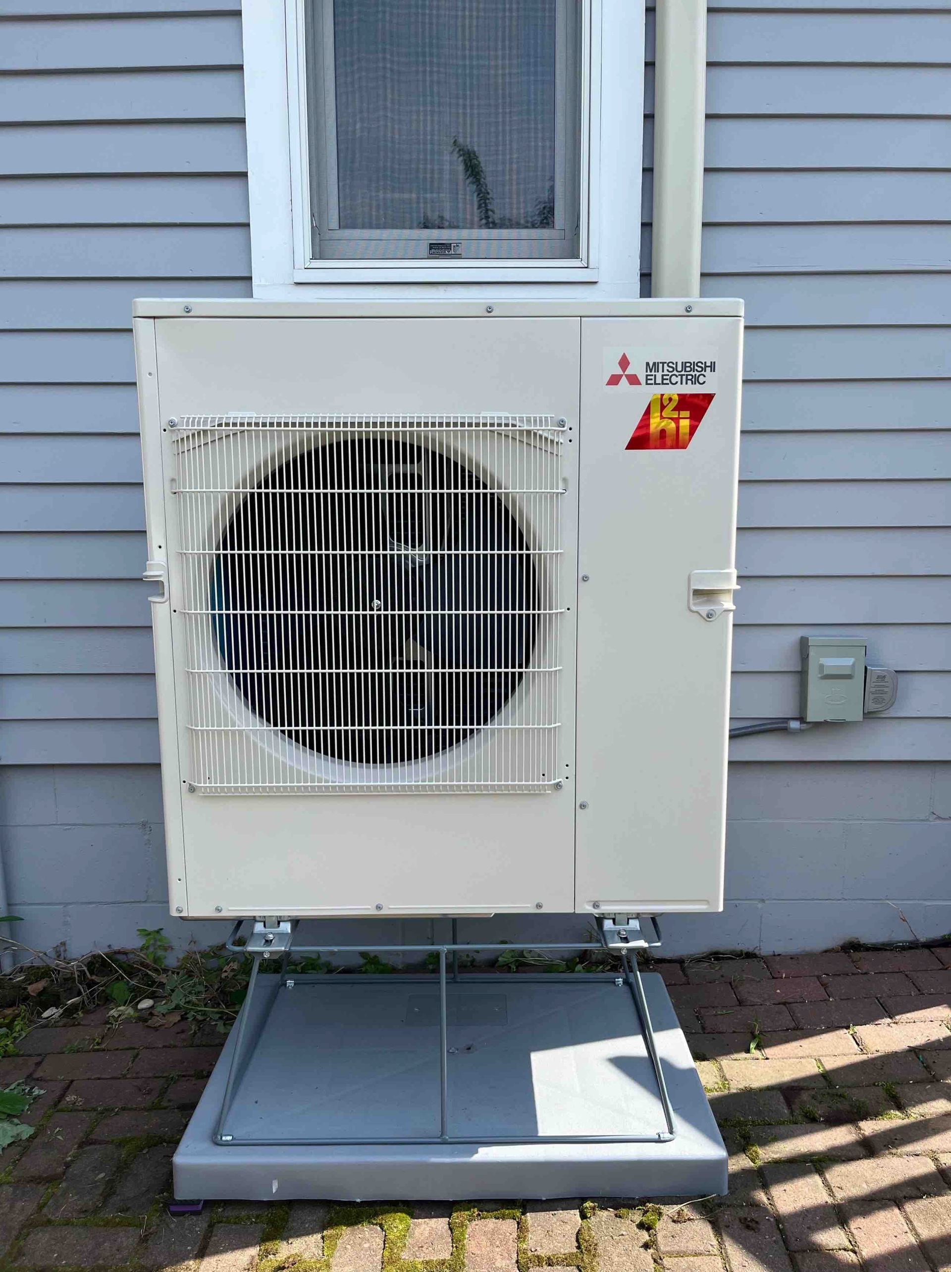 Mitsubishi heat pump unit mounted on a grey base next to a blue-grey house.