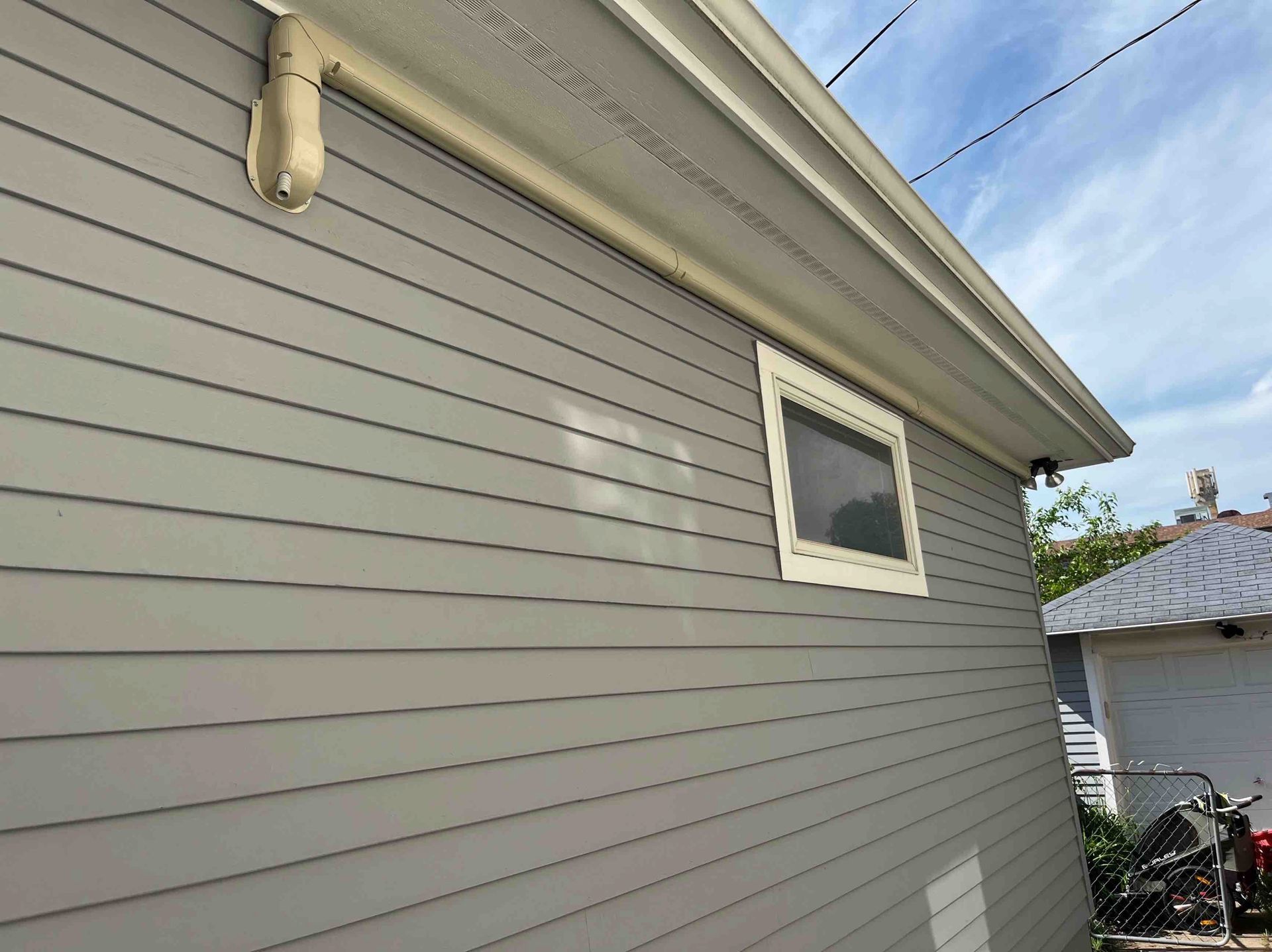 Gray-sided building with a window, white trim, and rain gutter against a blue sky.