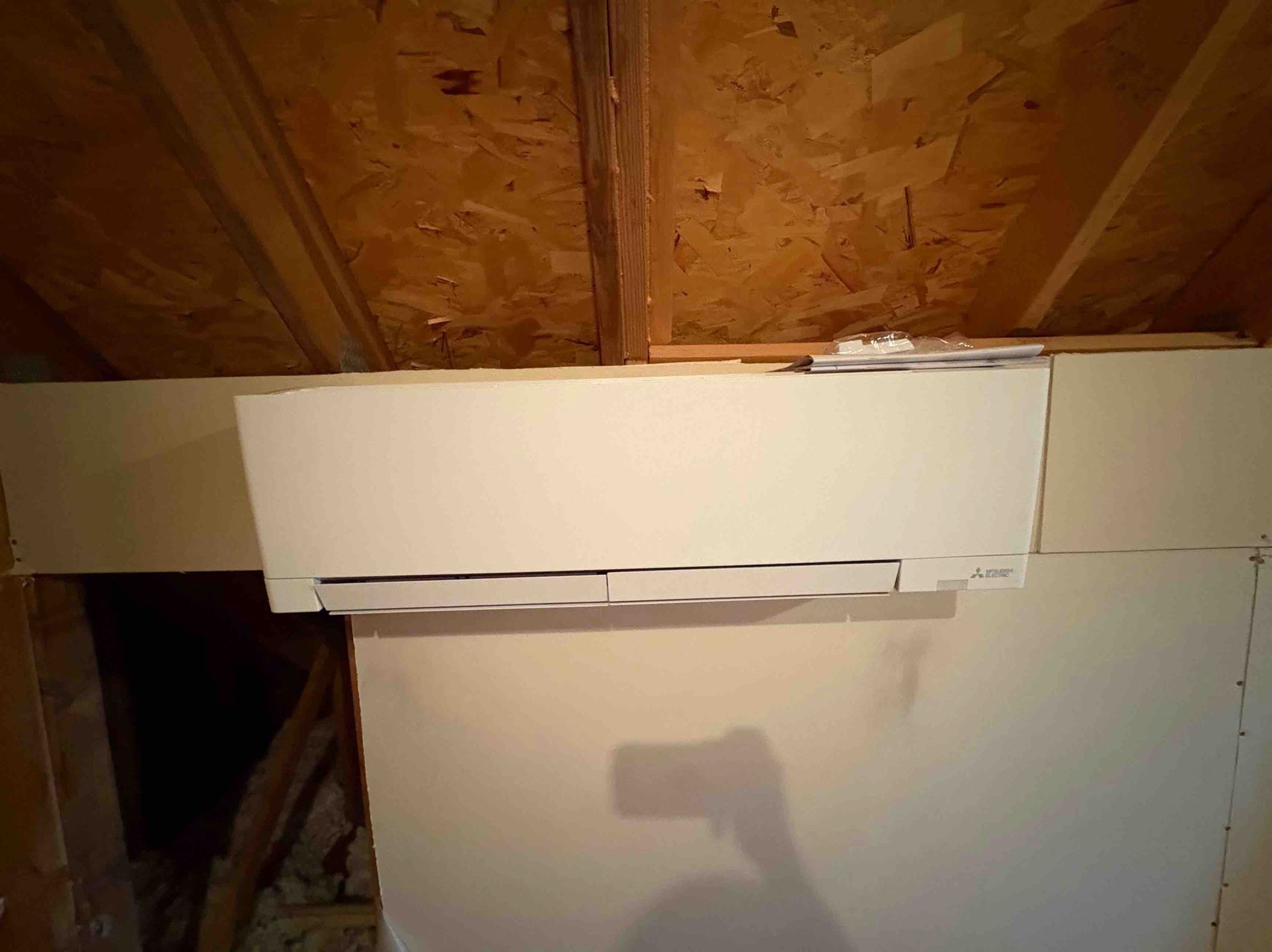 White wall-mounted air conditioning unit in an unfinished room, beneath a wooden beam.