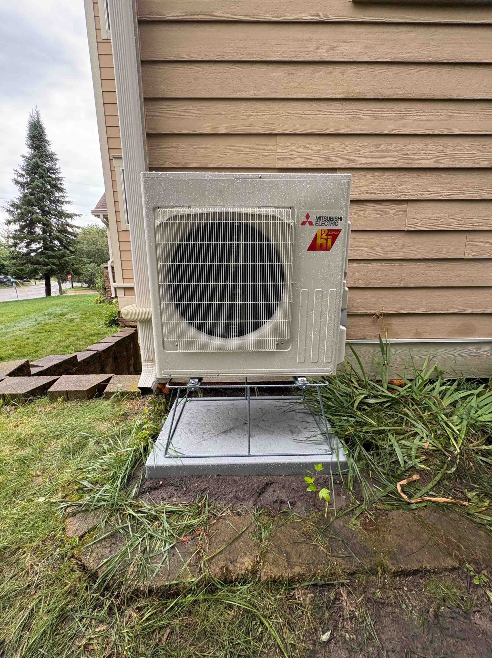 Mitsubishi AC unit mounted on a concrete pad, beside a house with tan siding.  Green grass surrounds it.