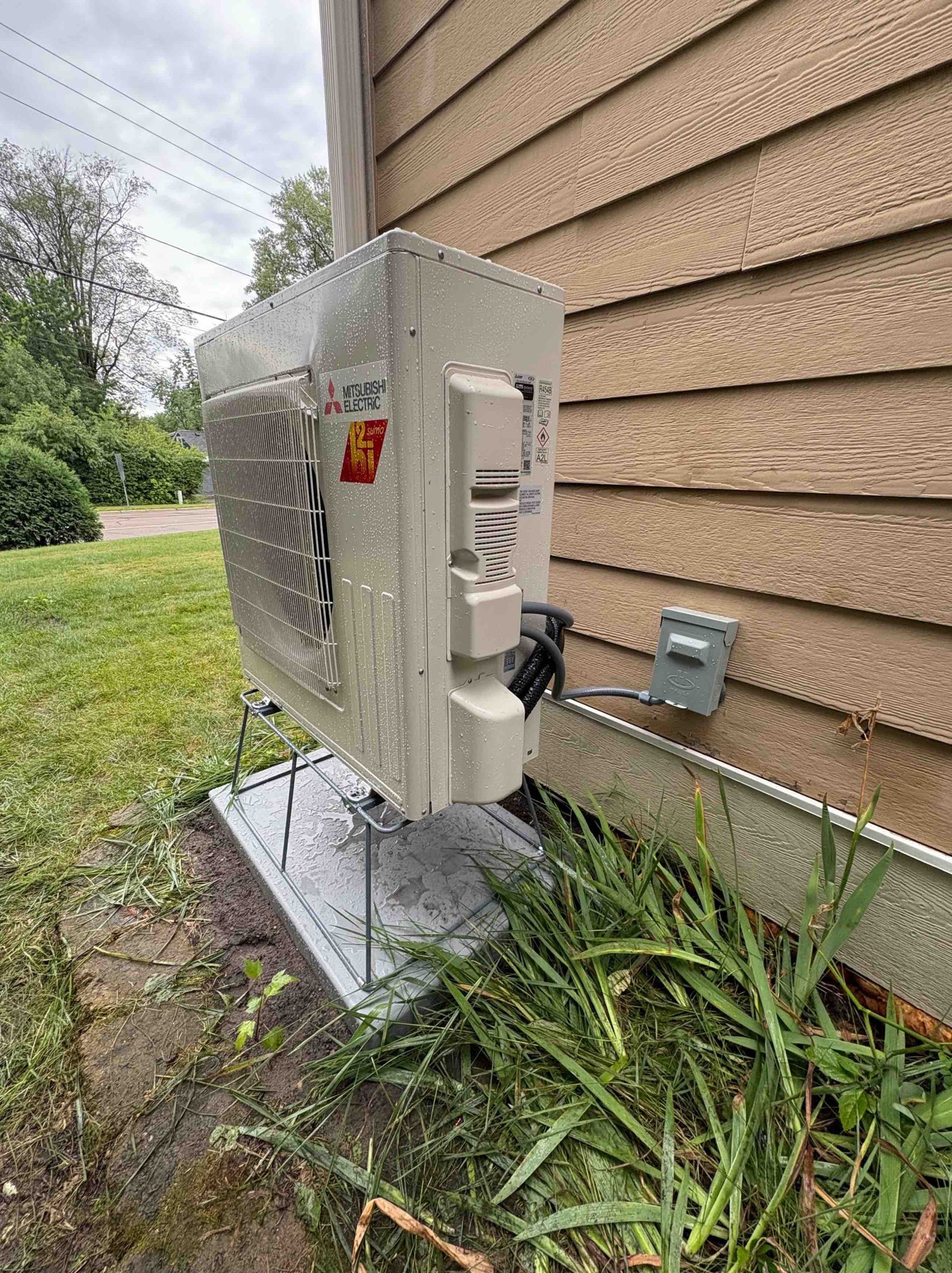 White Mitsubishi outdoor AC unit mounted on a metal base next to a wooden house.
