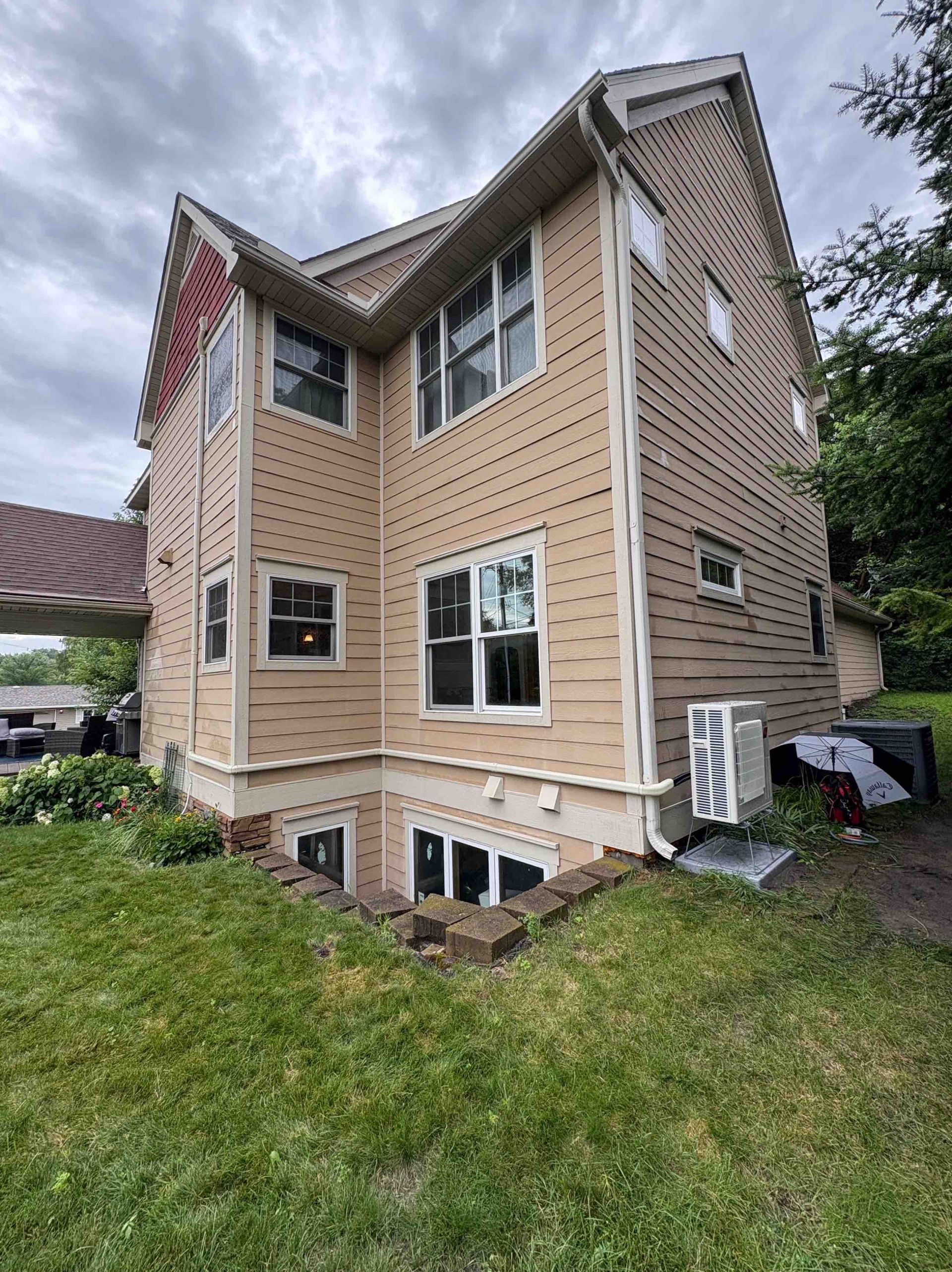 Tan house with several windows, nestled in green grass with a partial basement view and an air conditioning unit.