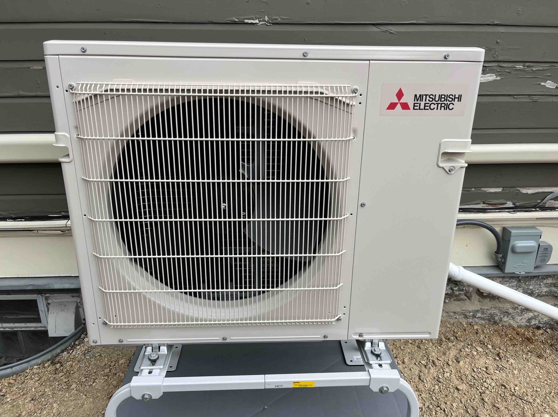 Mitsubishi mini-split heat pump mounted outdoors; metallic gray; fan behind a protective grille.