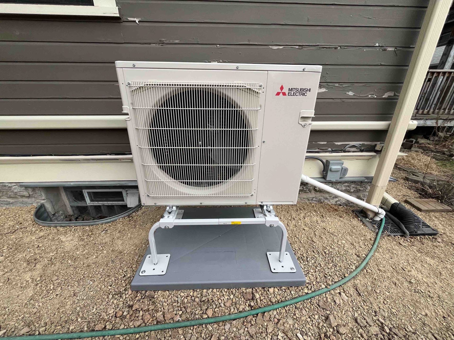 Mitsubishi heat pump, exterior view. Mounted on gray base, gravel ground, against brown siding.