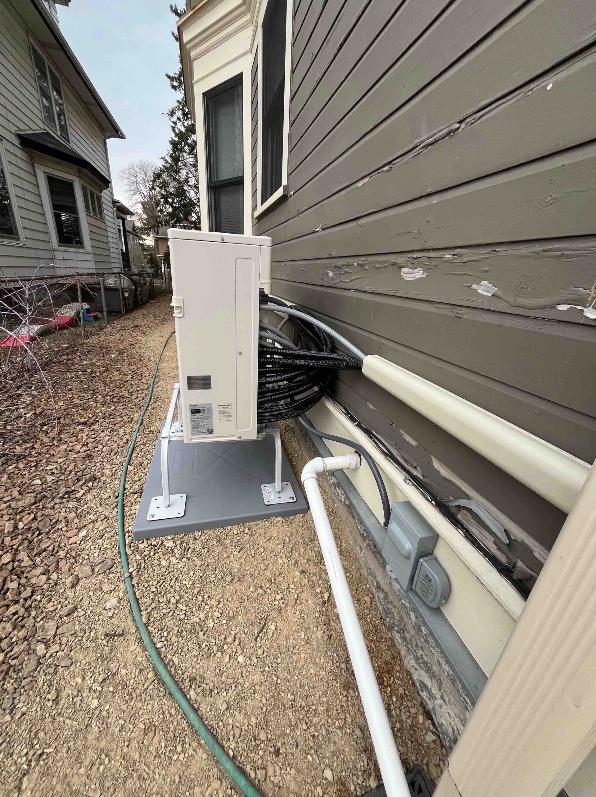 White box with wires attached to a gray house exterior, sitting on a gray platform, outdoors.