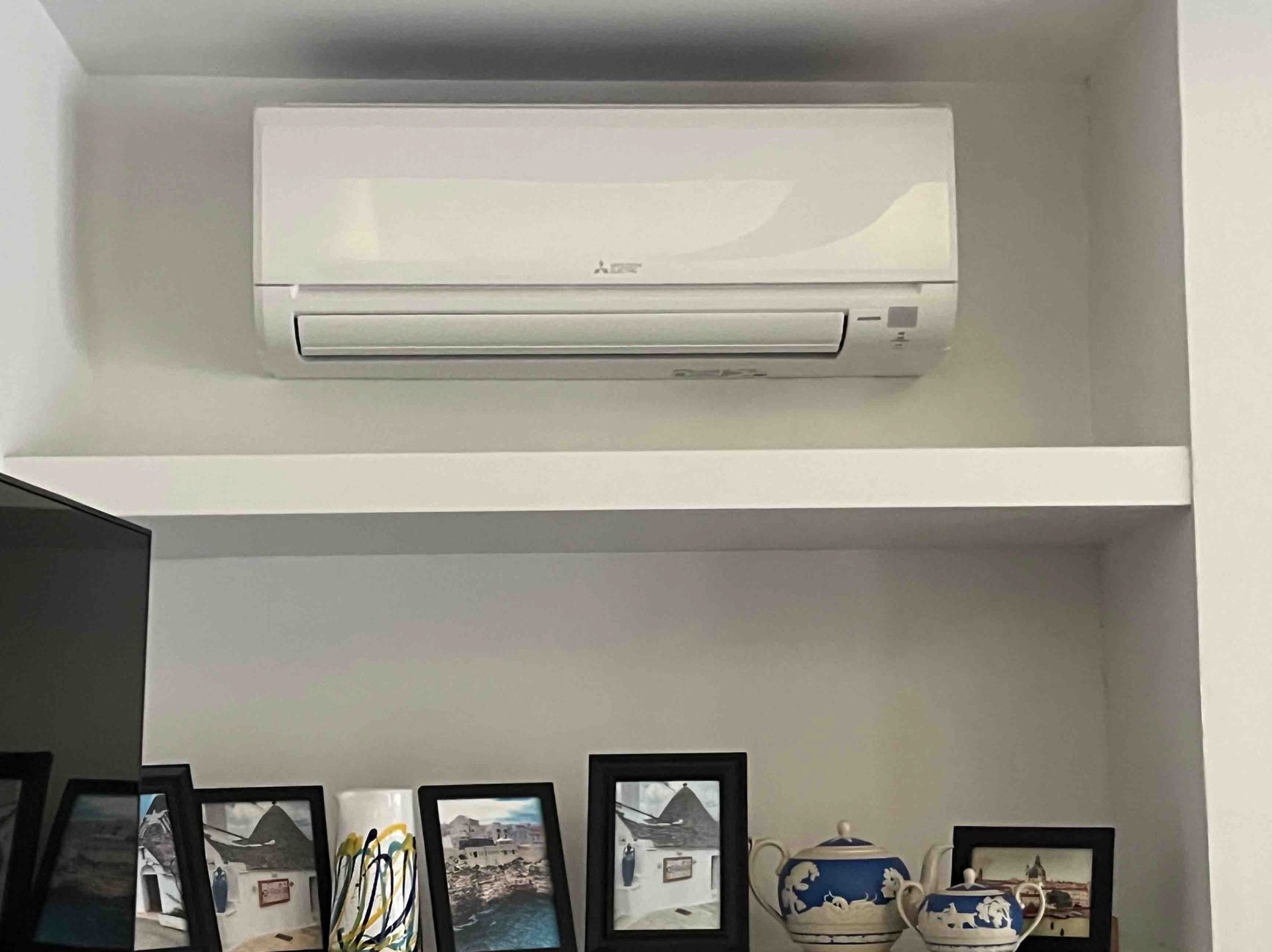 White air conditioner mounted above a shelf with framed photos and ceramic pieces.