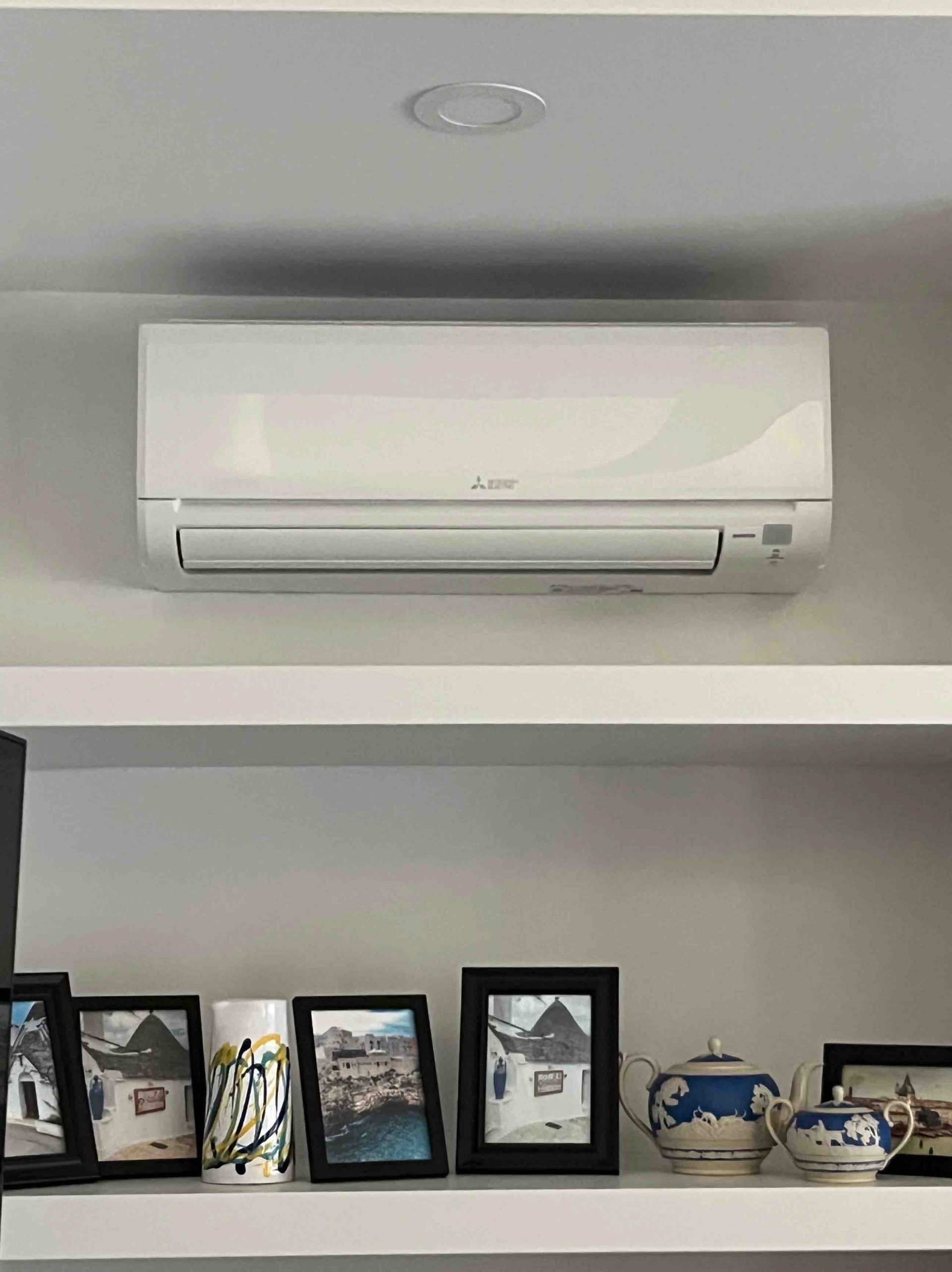 White air conditioner mounted above a shelf with framed photos and decorative items.