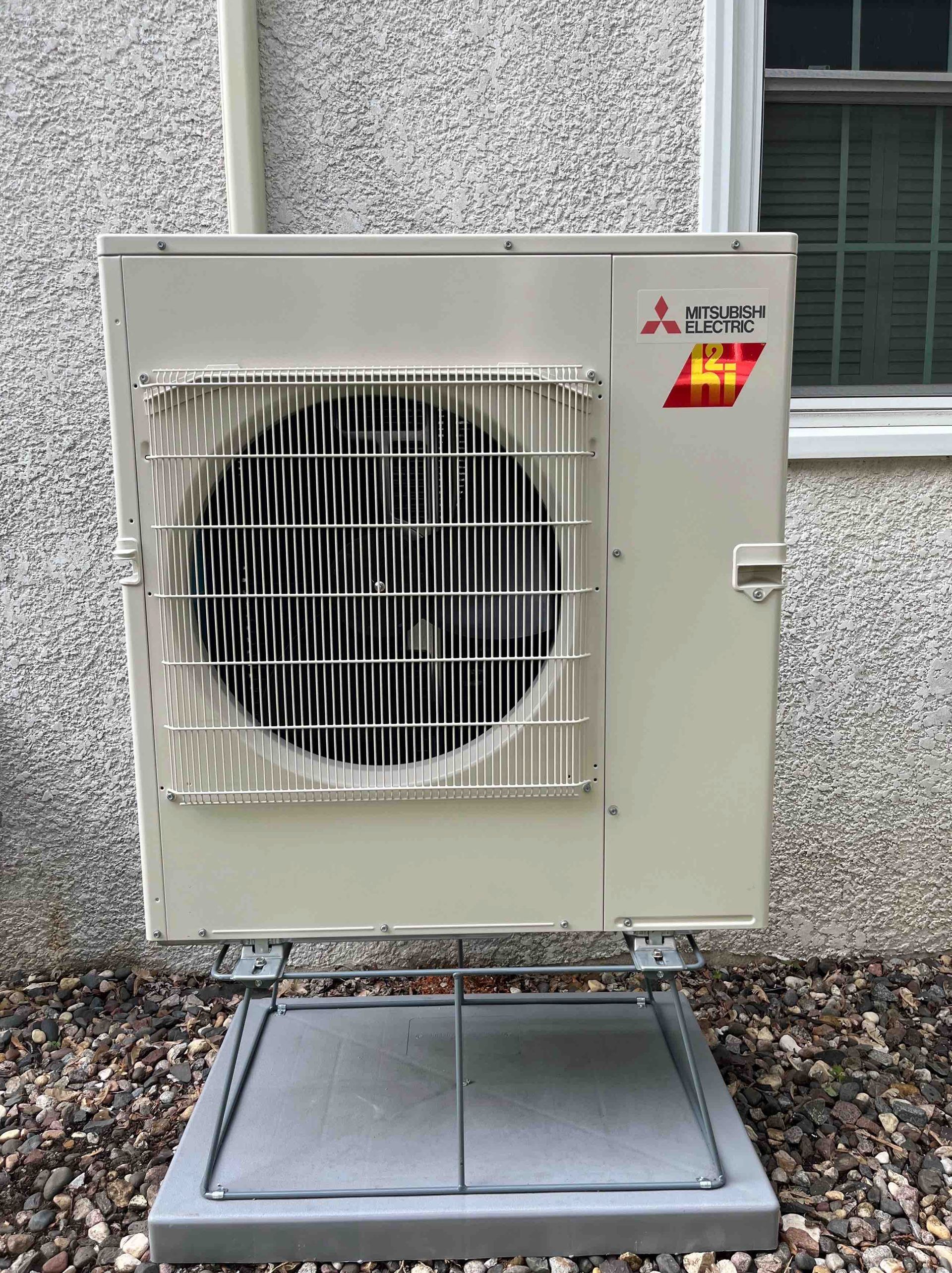 Mitsubishi heat pump unit on gravel, next to a wall and window.